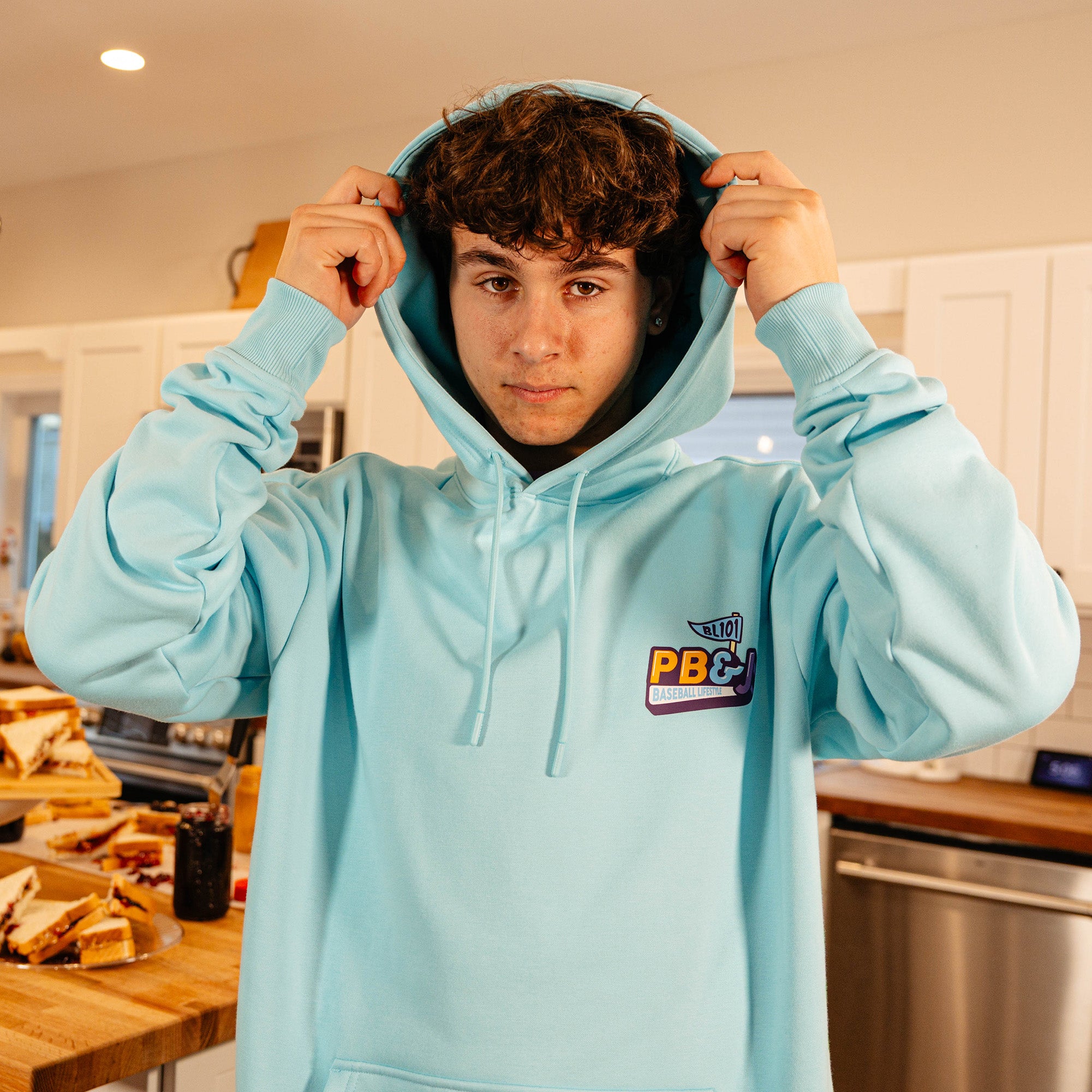 Male model pulling up the hood of the BL101 PB&J Baseball Lifestyle light blue hoodie in a kitchen, showing the chest logo.
