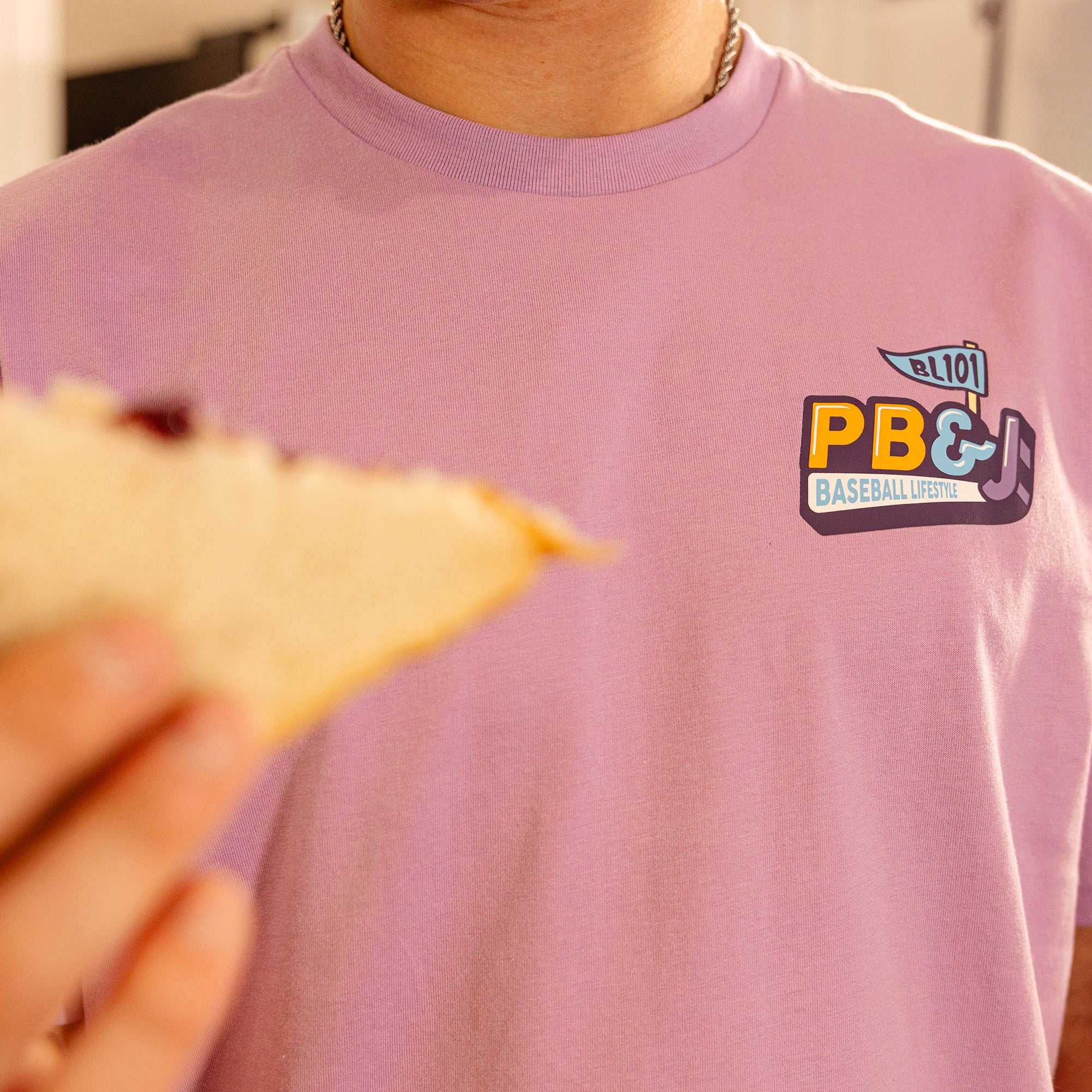 Close-up of the PB&J Baseball Lifestyle chest logo on the purple tee, with a PB&J sandwich in the foreground.