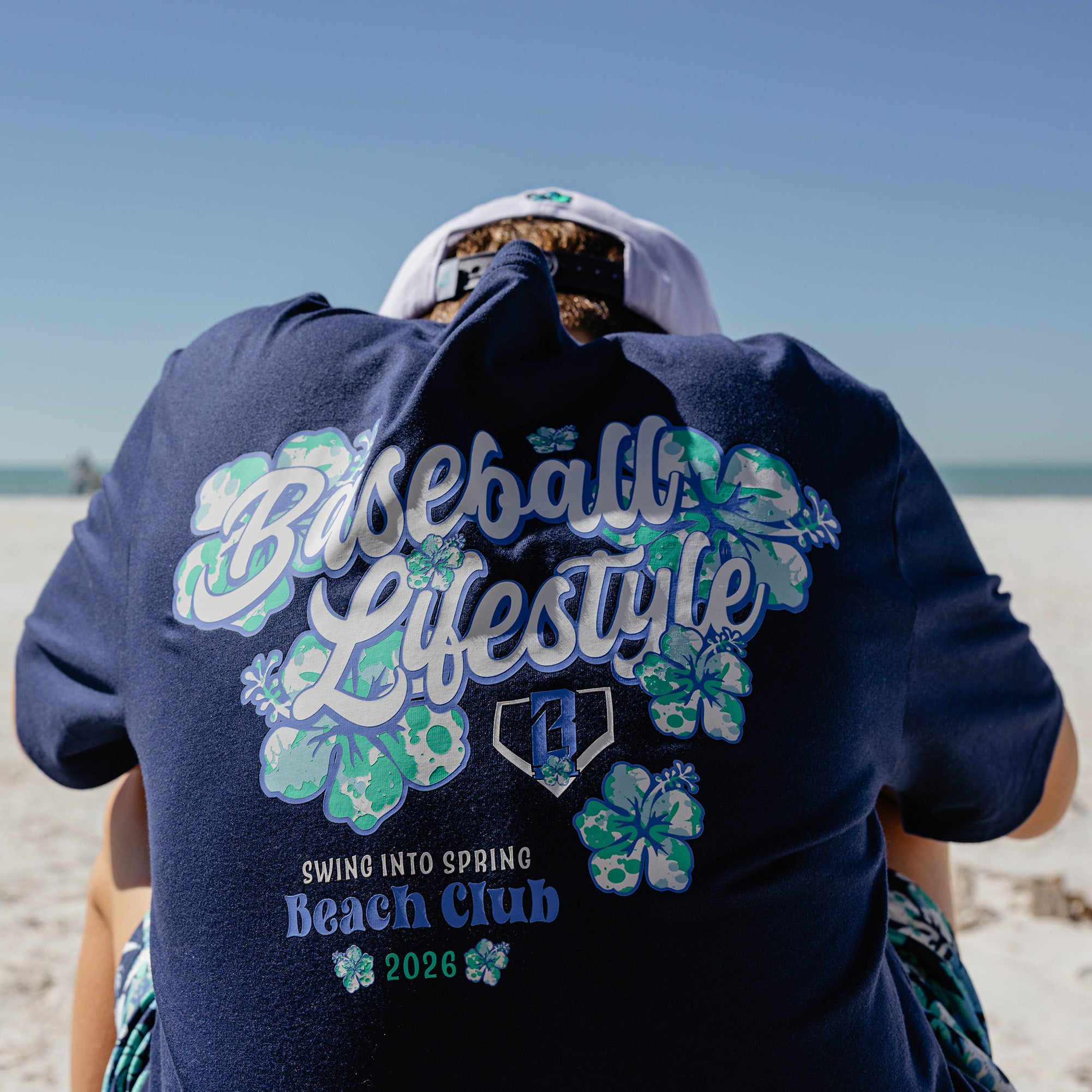 Person wearing a navy blue shirt with 'Baseball Lifestyle Swing into Spring Beach Club 2026' text and floral designs on a beach