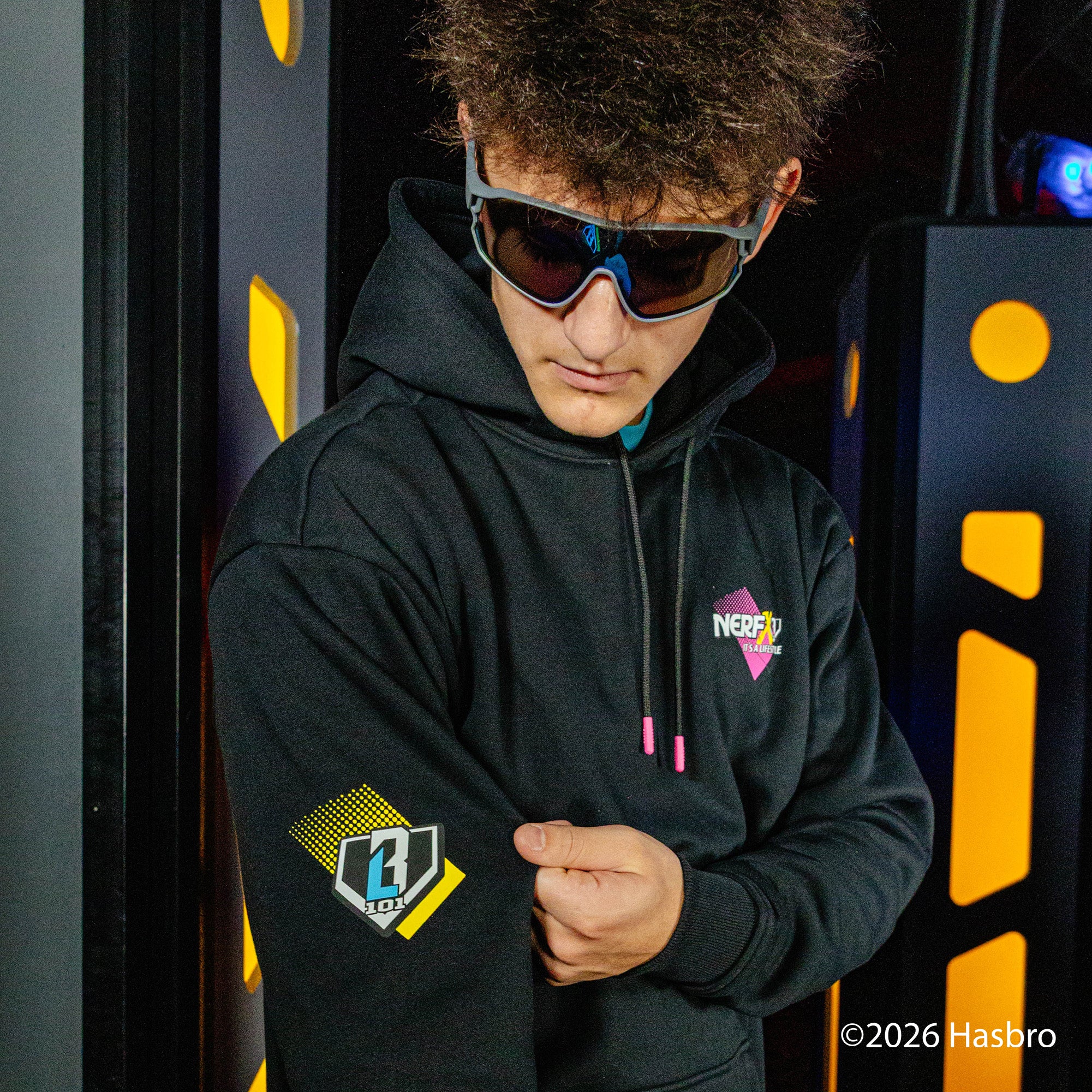Person wearing a black hoodie with NERF X BL101 logos, standing in front of a dark background with yellow accents.