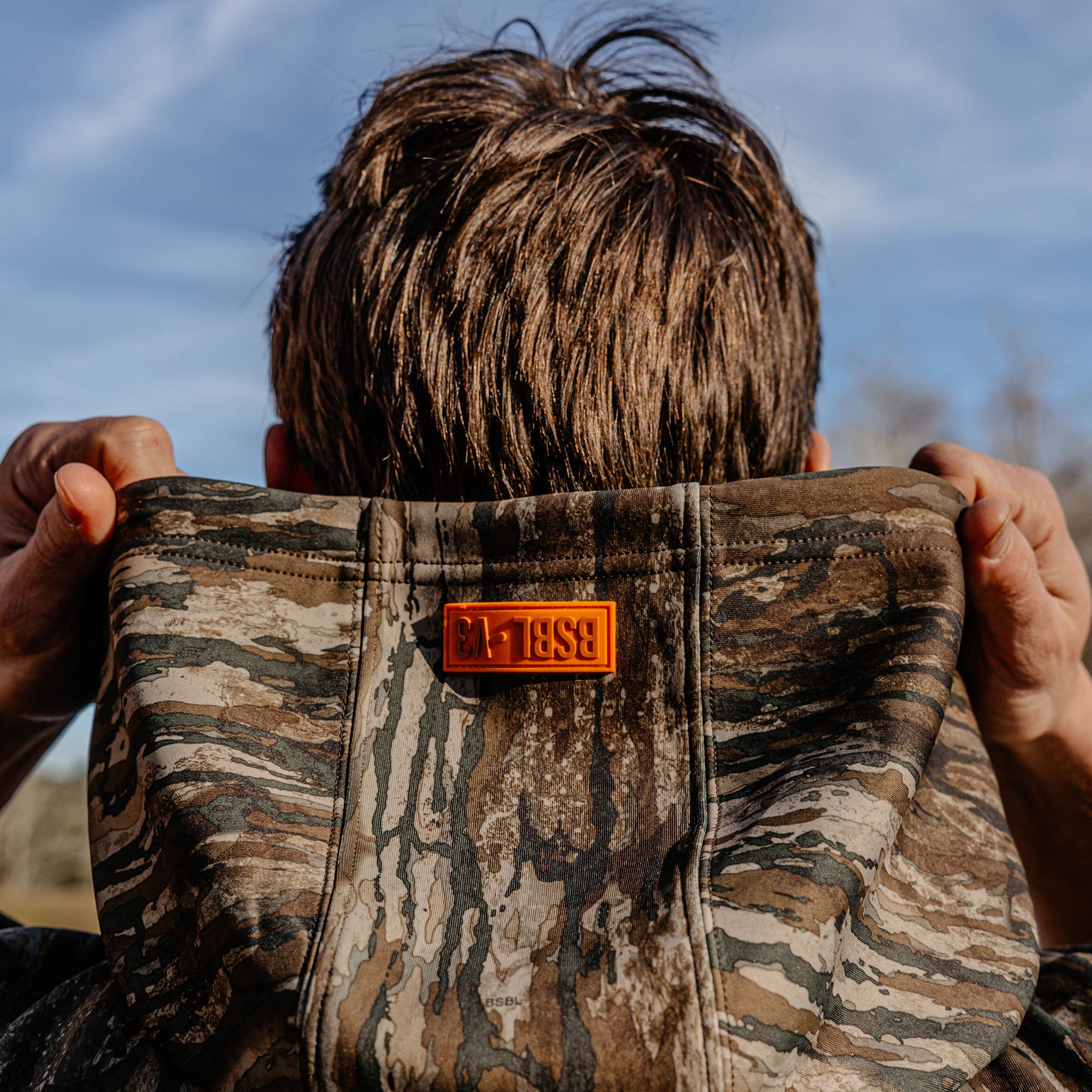 Person wearing a camouflage jacket with a 'BSBL V3' logo, standing outdoors.