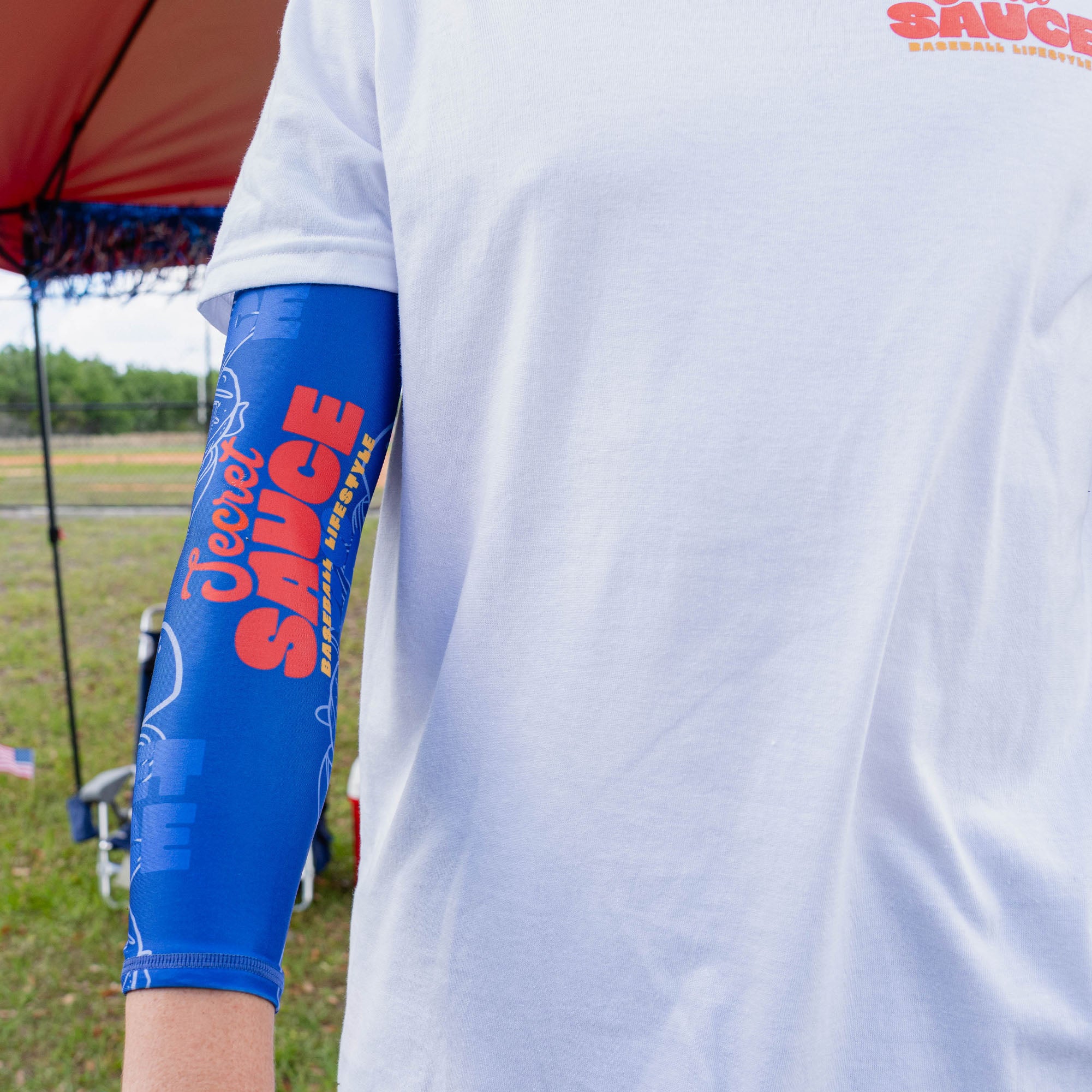 Side close-up of adult arm showing full blue Secret Sauce arm sleeve with Secret Sauce Baseball Lifestyle logo and burger line art pattern while wearing white Secret Sauce tee