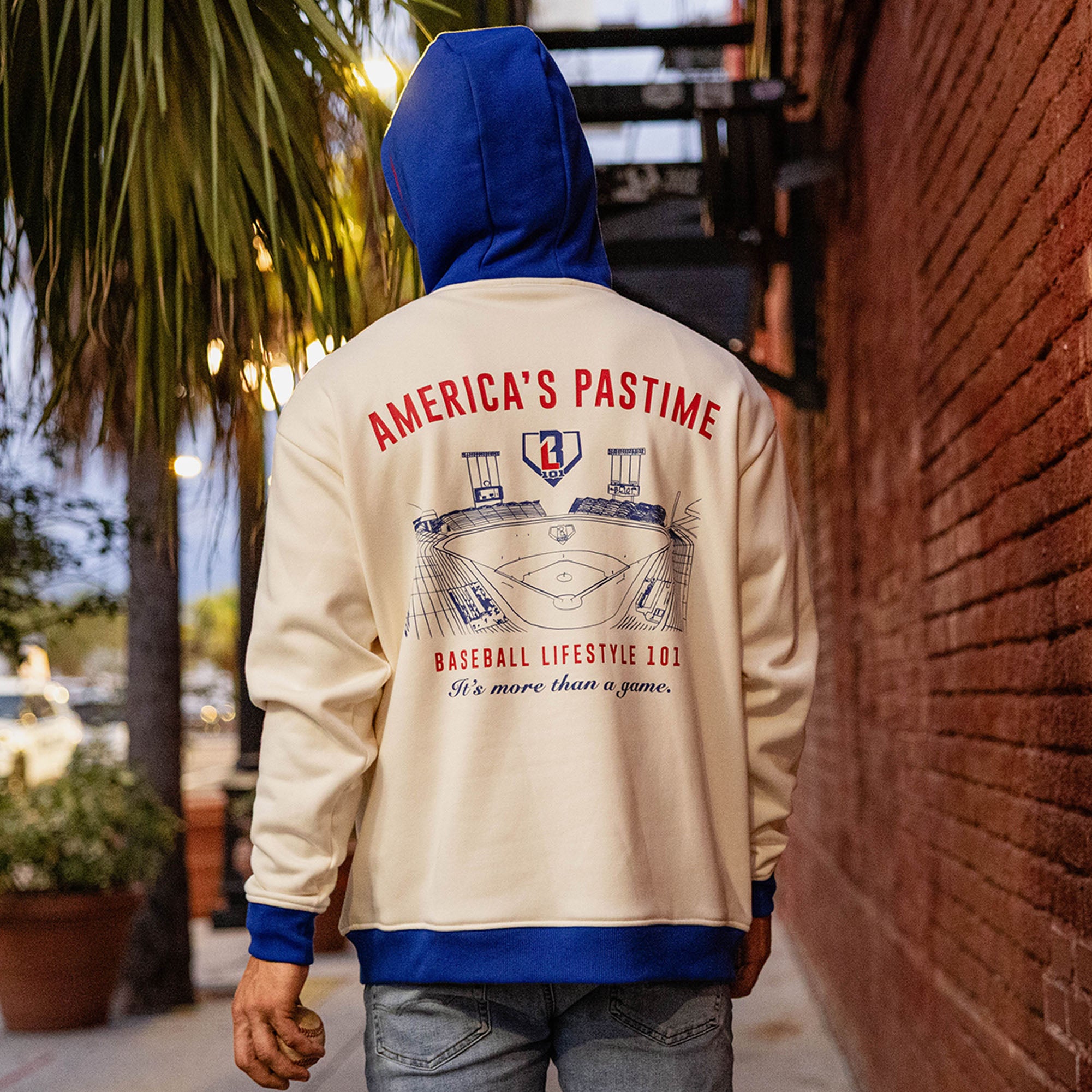 Back view of man on a sidewalk wearing a cream stadium hoodie with royal blue hood