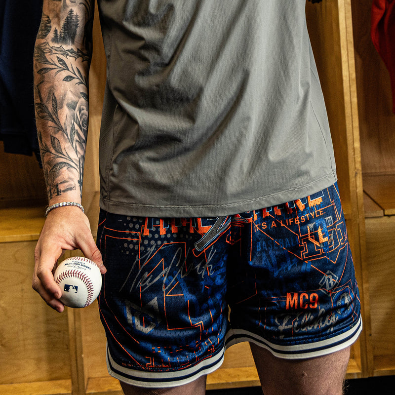 Person wearing blue athletic shorts with orange text and patterns, holding a baseball in a locker room.