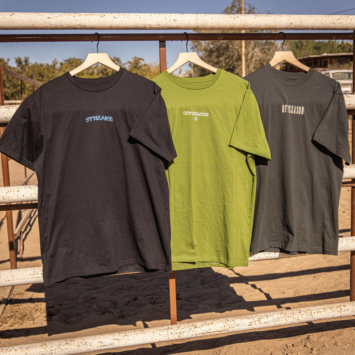 Three 'OFFSEASON' t-shirts in black, green, and gray hanging on a metal railing outdoors.