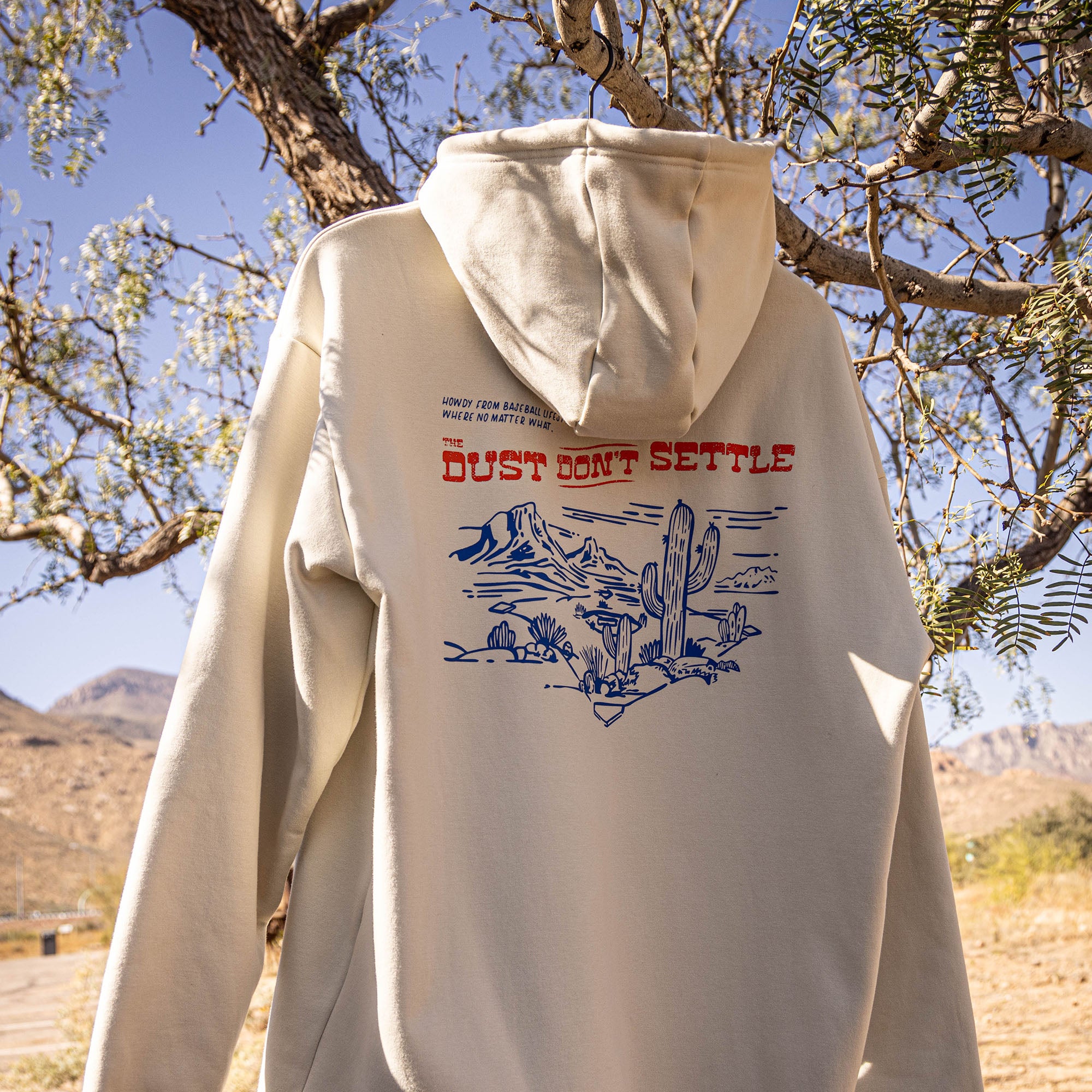 Beige hoodie with desert graphic and text 'Dust Don't Settle' on back with a desert landscape in the background.