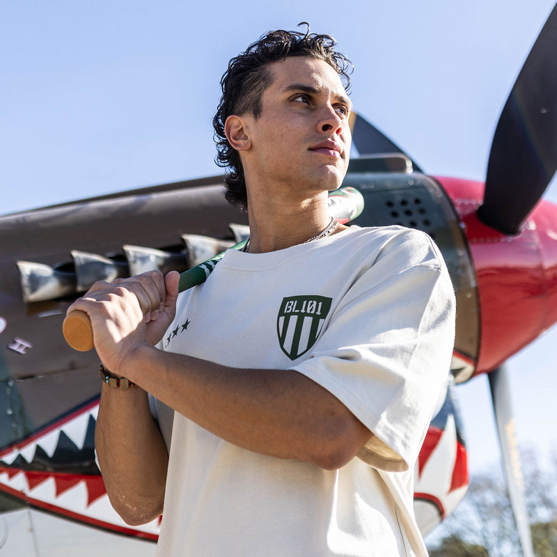 Person holding a baseball bat in front of a red airplane wearing a Tan t-shirt with dark green graphic with 4 stars and armor BL101 logo