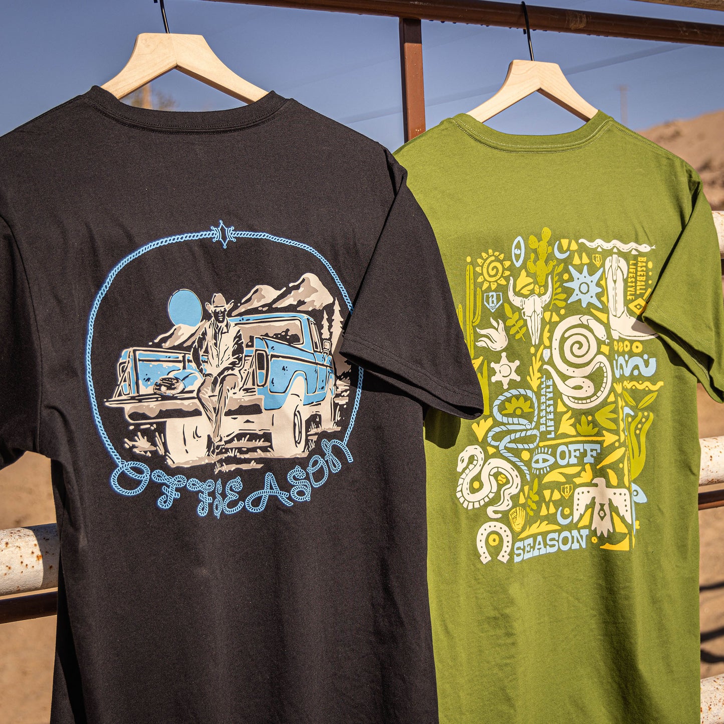 Two t-shirts, green and black,  with 'OFFSEASON' desert graphic designs on a wooden rack outdoors.