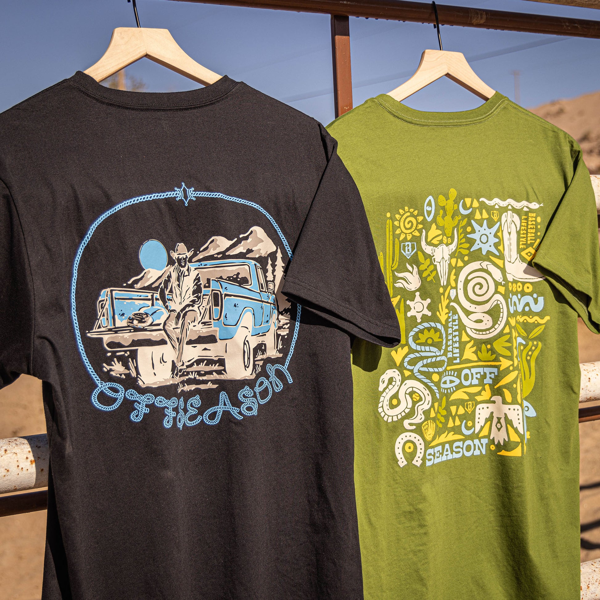 Two t-shirts, green and black,  with 'OFFSEASON' desert graphic designs on a wooden rack outdoors.
