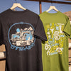 Two t-shirts, green and black,  with 'OFFSEASON' desert graphic designs on a wooden rack outdoors.