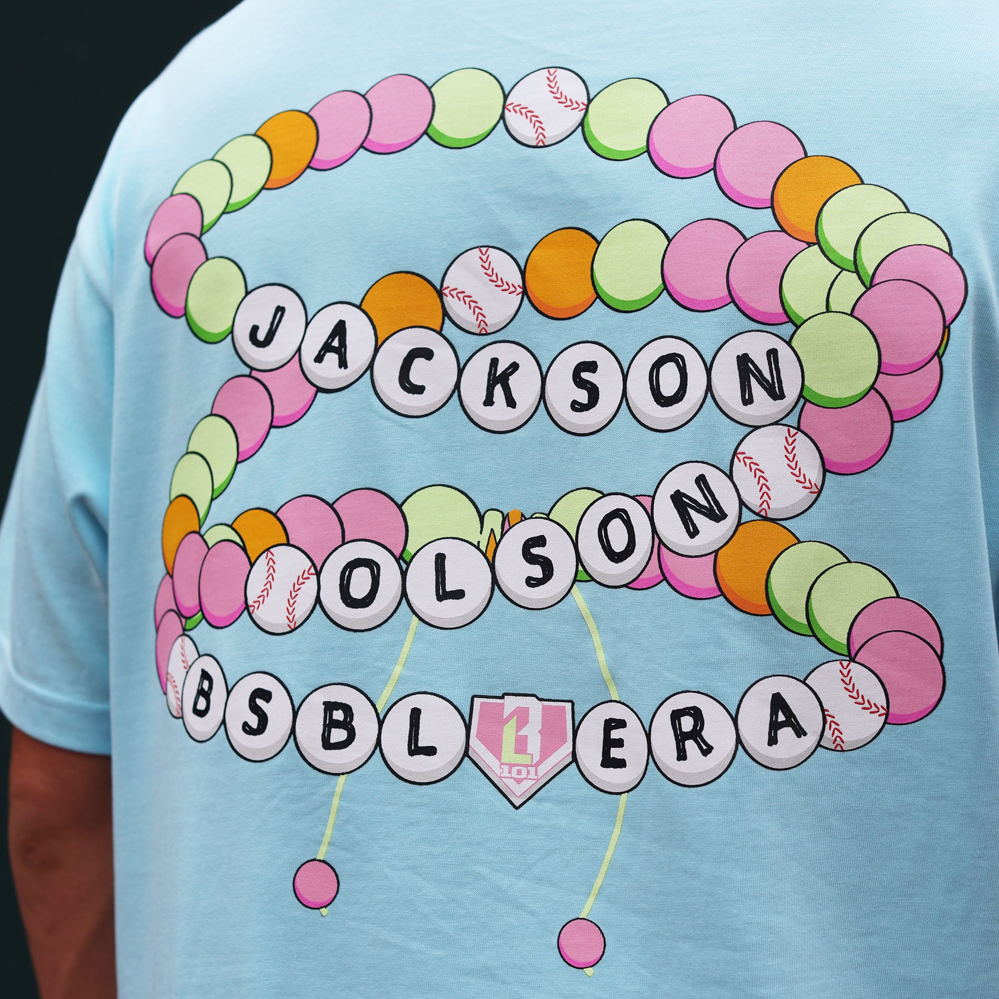Close-up of the back graphic on the light blue BSBL Era tee, featuring a colorful friendship bracelet design with "Jackson Olson BSBL Era" lettering and baseball bead details