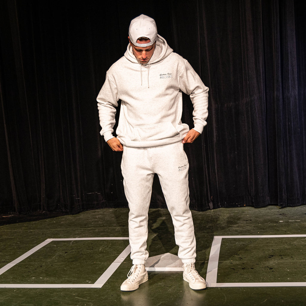 Adult male model looking down while wearing the heather gray Jackson Olson x BL101 BSBL Era sweatpants and matching hoodie, showing the full coordinated set