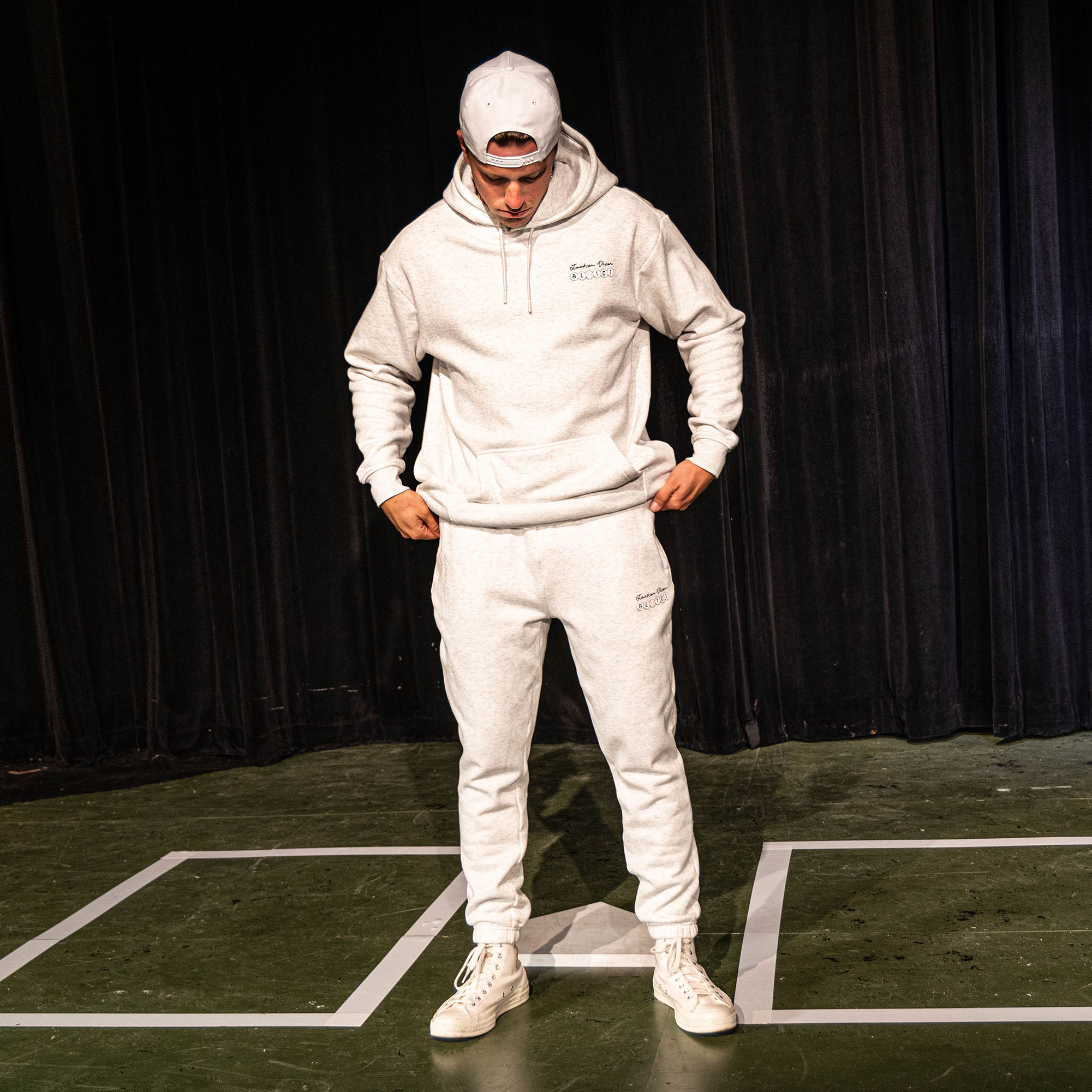 Adult male model looking down while wearing the heather gray Jackson Olson x BL101 BSBL Era sweatpants and matching hoodie, showing the full coordinated set