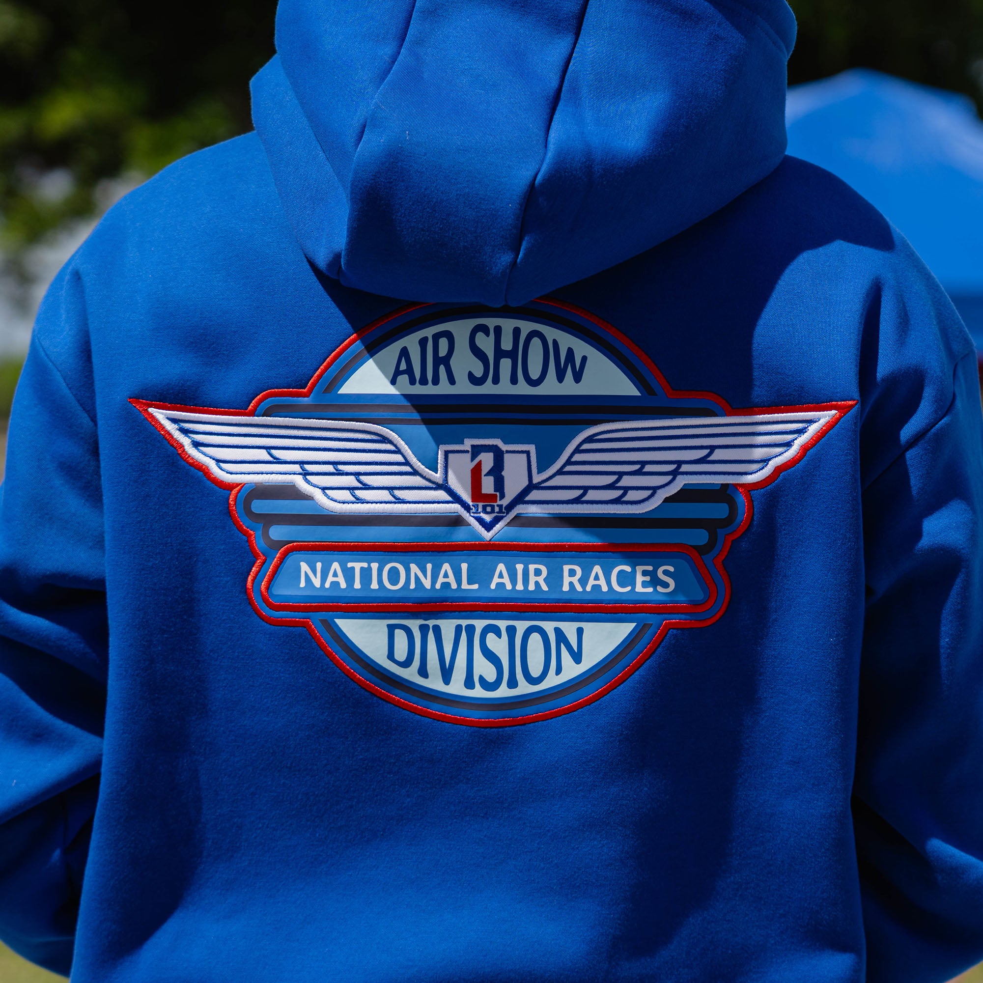 Close-up back detail of adult male model showing Air Show National Air Races Division embroidered badge on royal blue hoodie outdoors