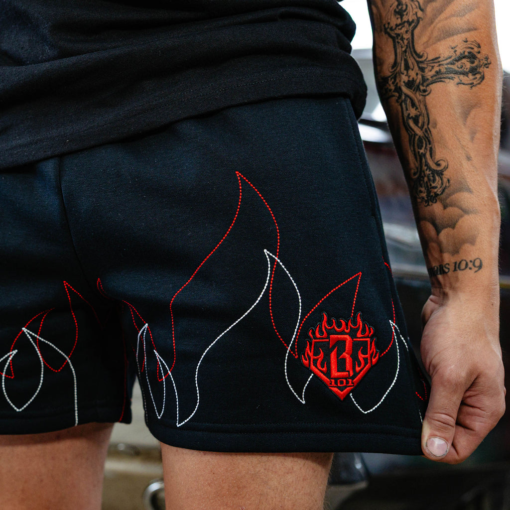 Close-up of stitched red and white flame graphic and embroidered red BL101 diamond logo on black sweatshorts, garage setting.