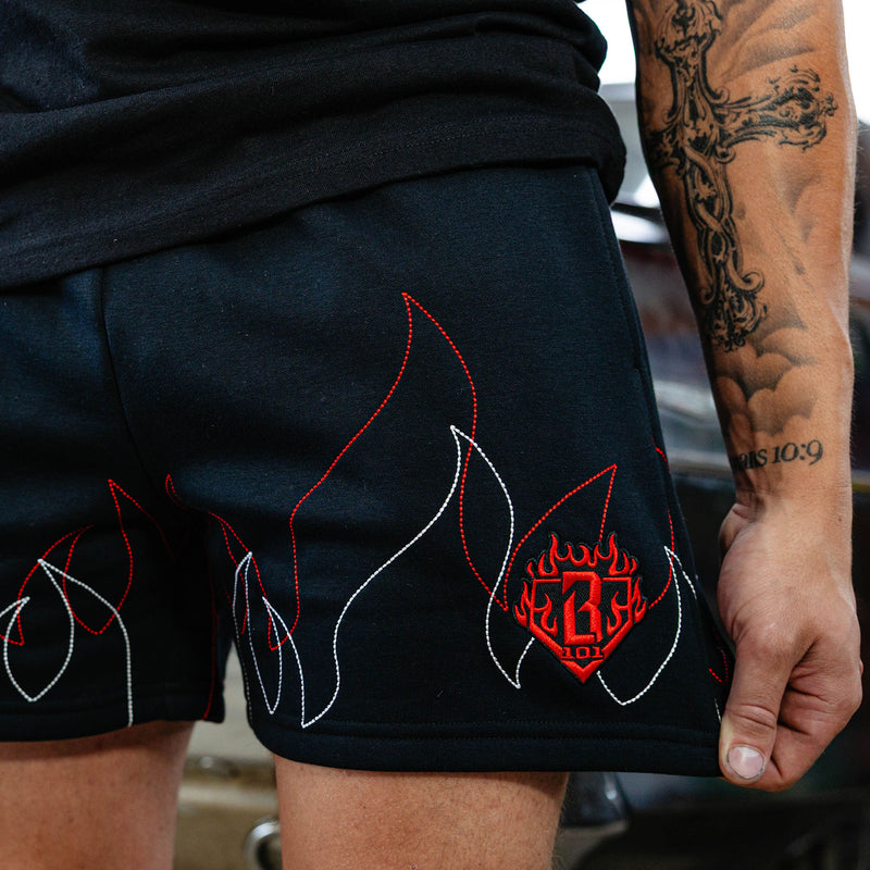 Close-up of stitched red and white flame graphic and embroidered red BL101 diamond logo on black sweatshorts, garage setting.