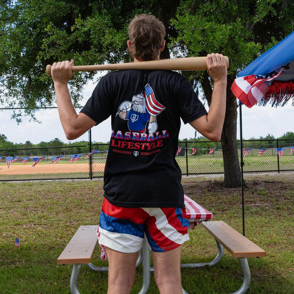 Back view of adult male model in red white and blue Patriot Pitch shorts and black Patriot Pitch tee holding baseball bat across shoulders at outdoor baseball event