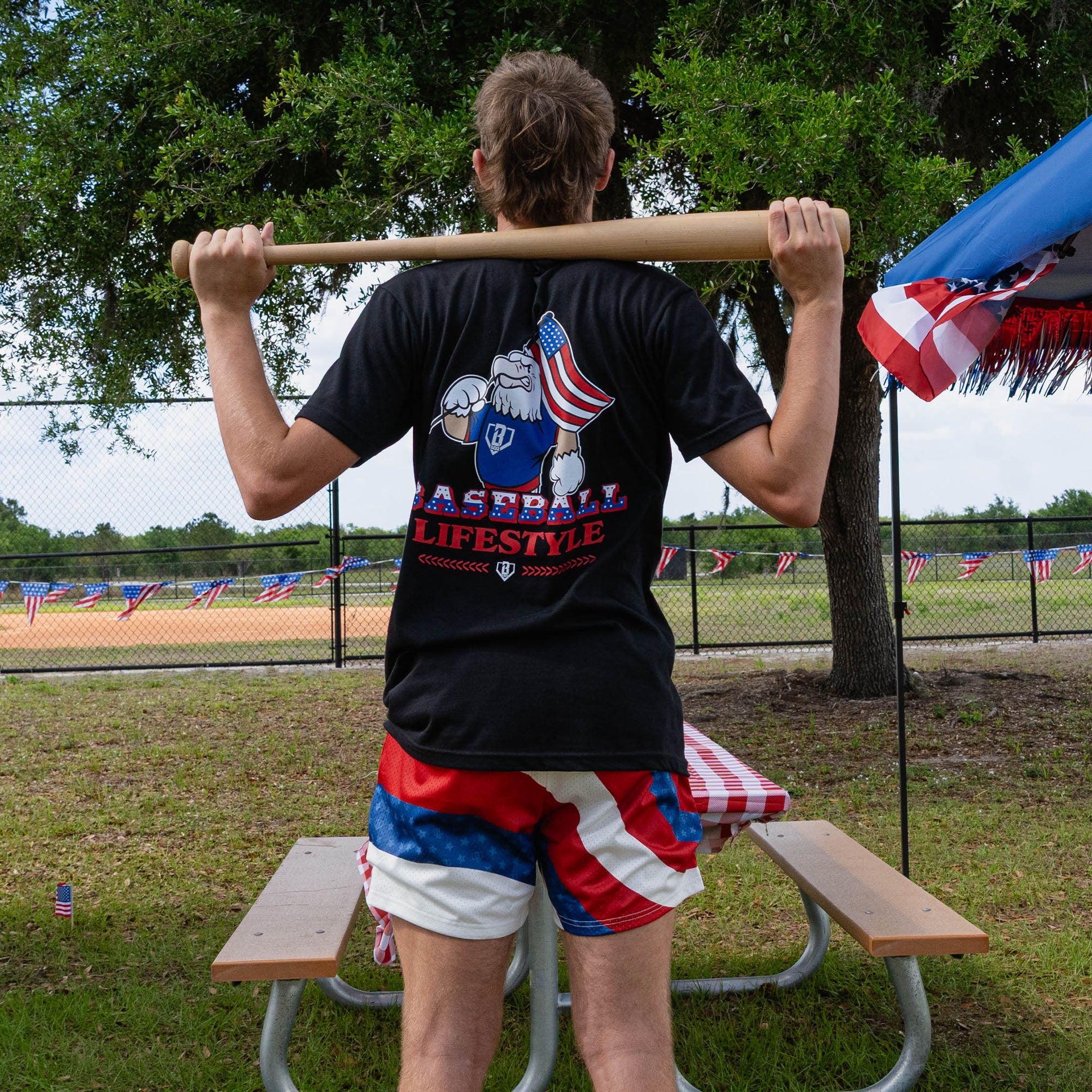 Back view of adult male model in red white and blue Patriot Pitch shorts and black Patriot Pitch tee holding baseball bat across shoulders at outdoor baseball event