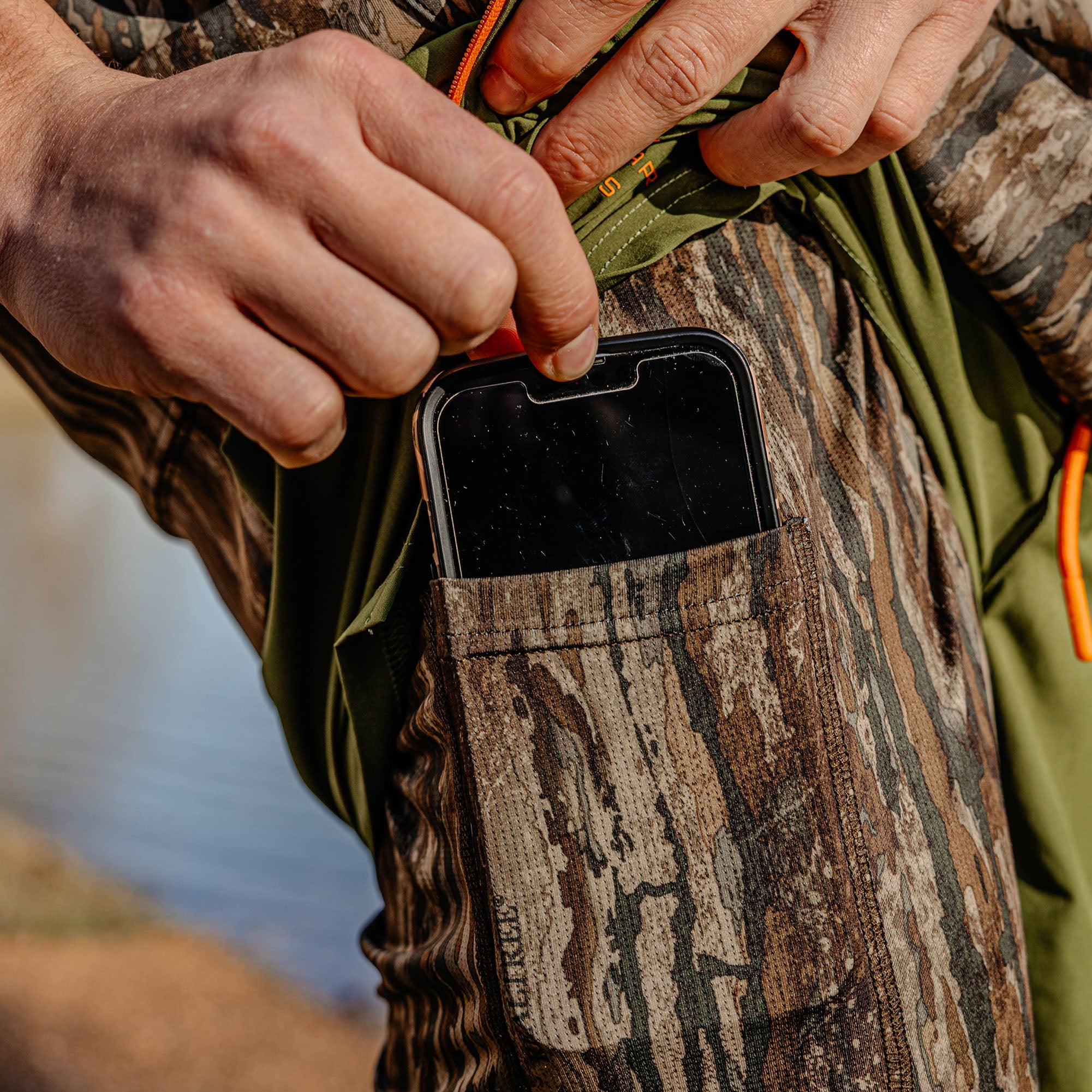 Person in camouflage clothing using a phone case with a natural setting in the background