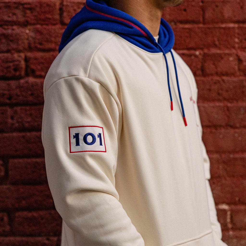 Close up of cream stadium hoodie sleeve showing embroidered 101 patch and royal blue cuff against a brick wall