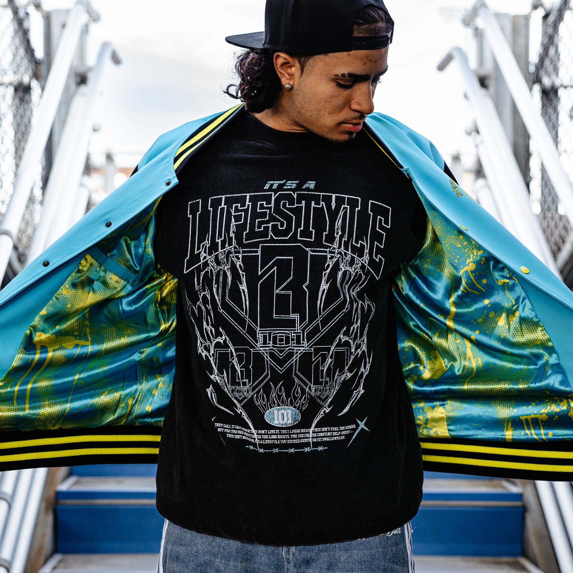 Person wearing a black t-shirt with graphic design and text, and blue and yellow jacket standing against a colorful background.