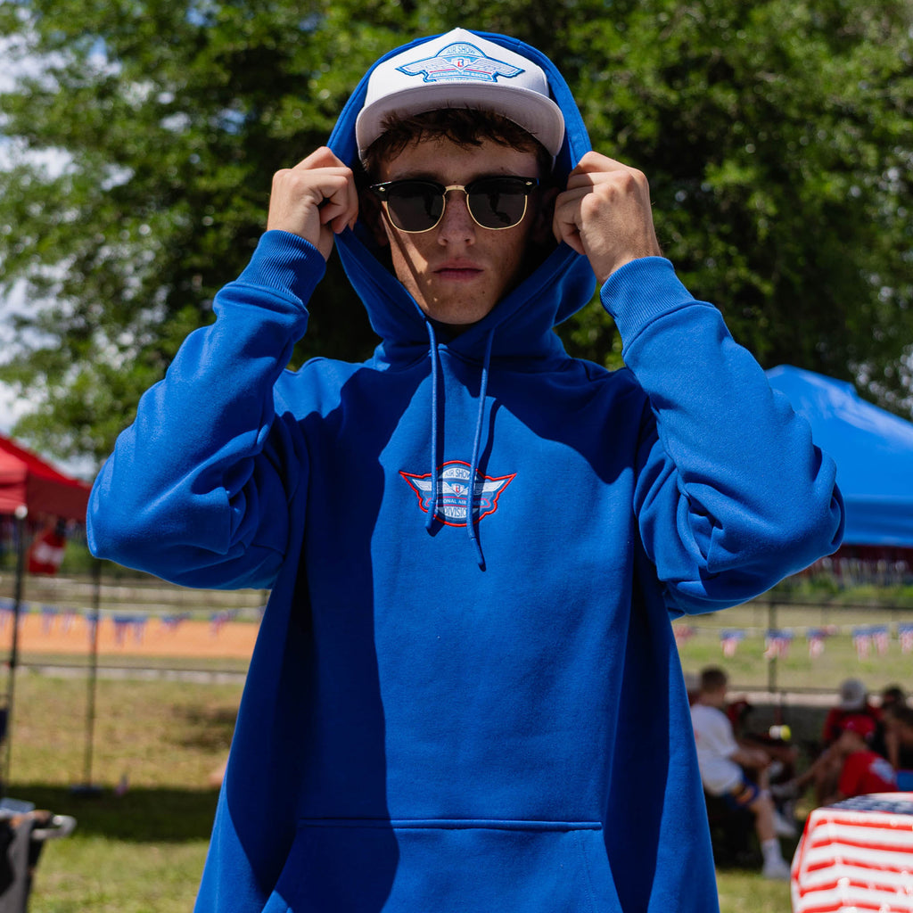 Adult male model in royal blue Fastball Flight hoodie pulling hood up over Fastball Flight snapback with sunglasses at outdoor baseball event