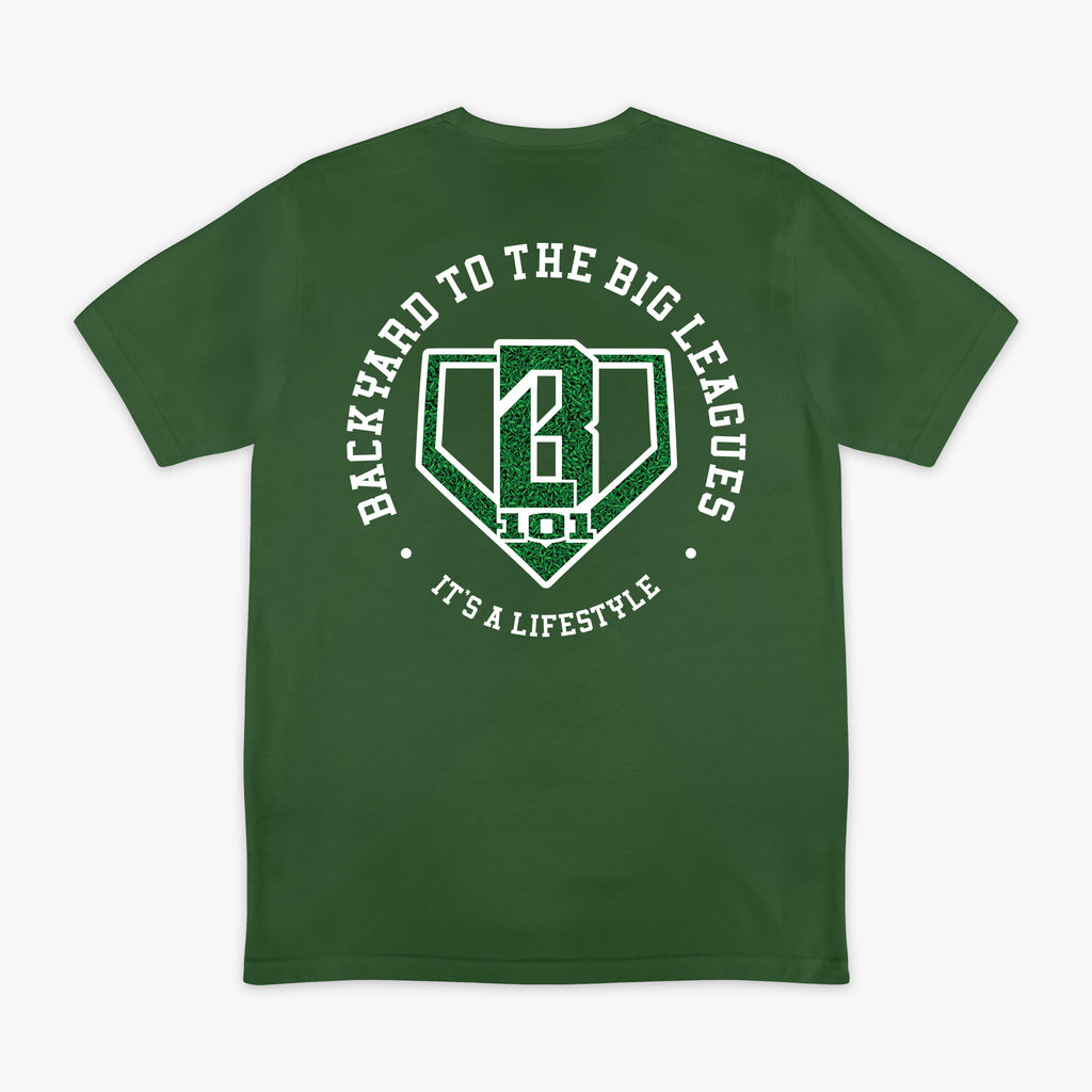 Image 1: Back flat lay of the BL101 Backyard to the Big Leagues forest green tee, featuring a circular "It's a Lifestyle" graphic with a grass-textured BL101 logo on a white background.
