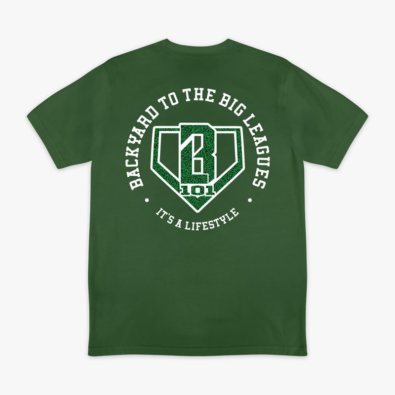 Image 1: Back flat lay of the BL101 Backyard to the Big Leagues forest green tee, featuring a circular "It's a Lifestyle" graphic with a grass-textured BL101 logo on a white background.