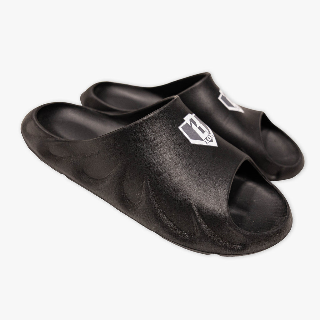 Pair of BL101 black slides on sand with the home plate logo visible on each shoe.