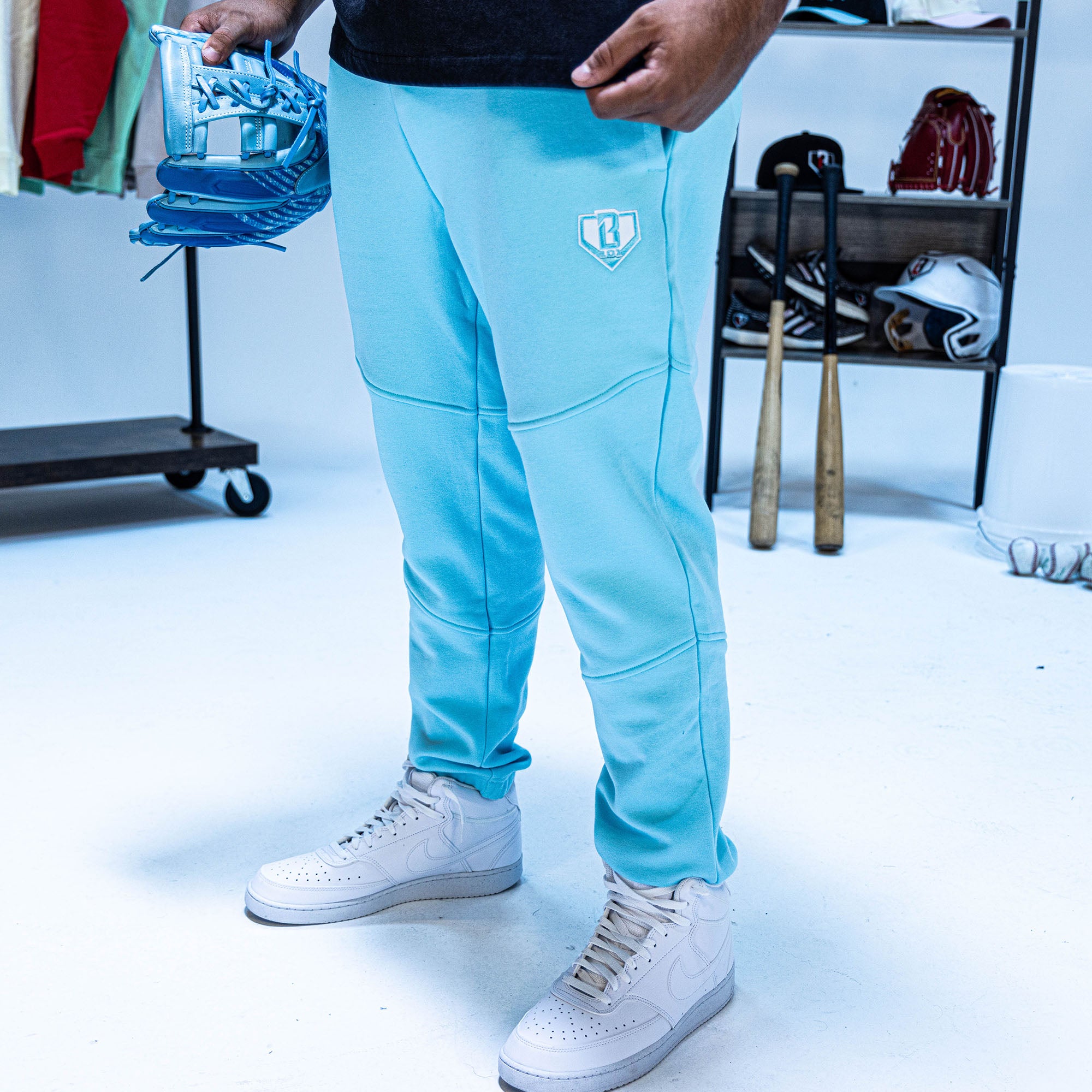 Person wearing light blue pants with a BL101 logo, white sneakers, holding blue shoes in a room with sports equipment.