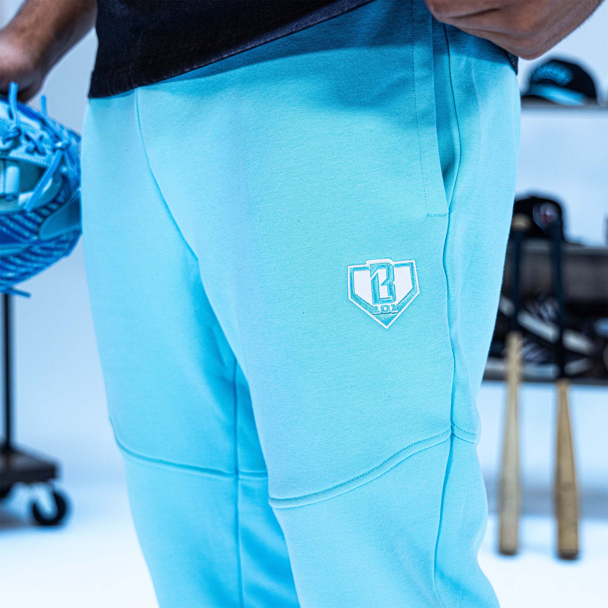 Light blue athletic pants with a BL101 logo on the back, worn by a person, with sports equipment in the background.