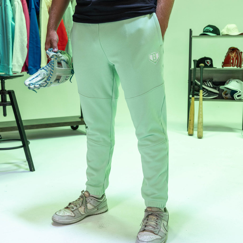 Person wearing light green jogger pants with a white BL101 logo, holding sports equipment in a room with sports equipment in the background.