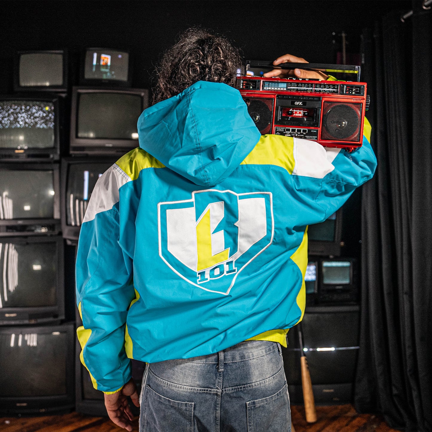 Person wearing a blue, yellow and white jacket with a BL101 logo, holding a boombox in front of vintage TVs.