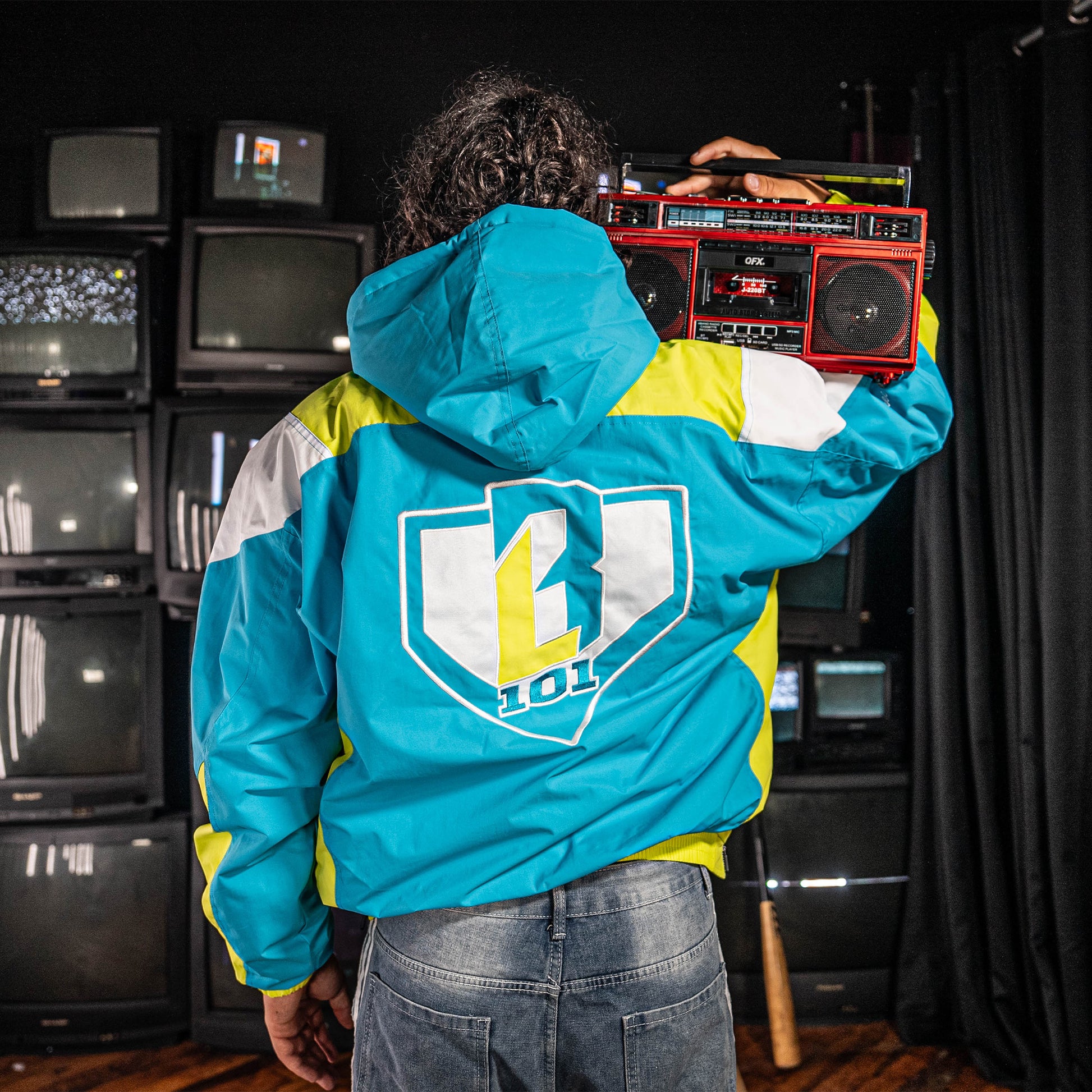 Person wearing a blue, yellow and white jacket with a BL101 logo, holding a boombox in front of vintage TVs.