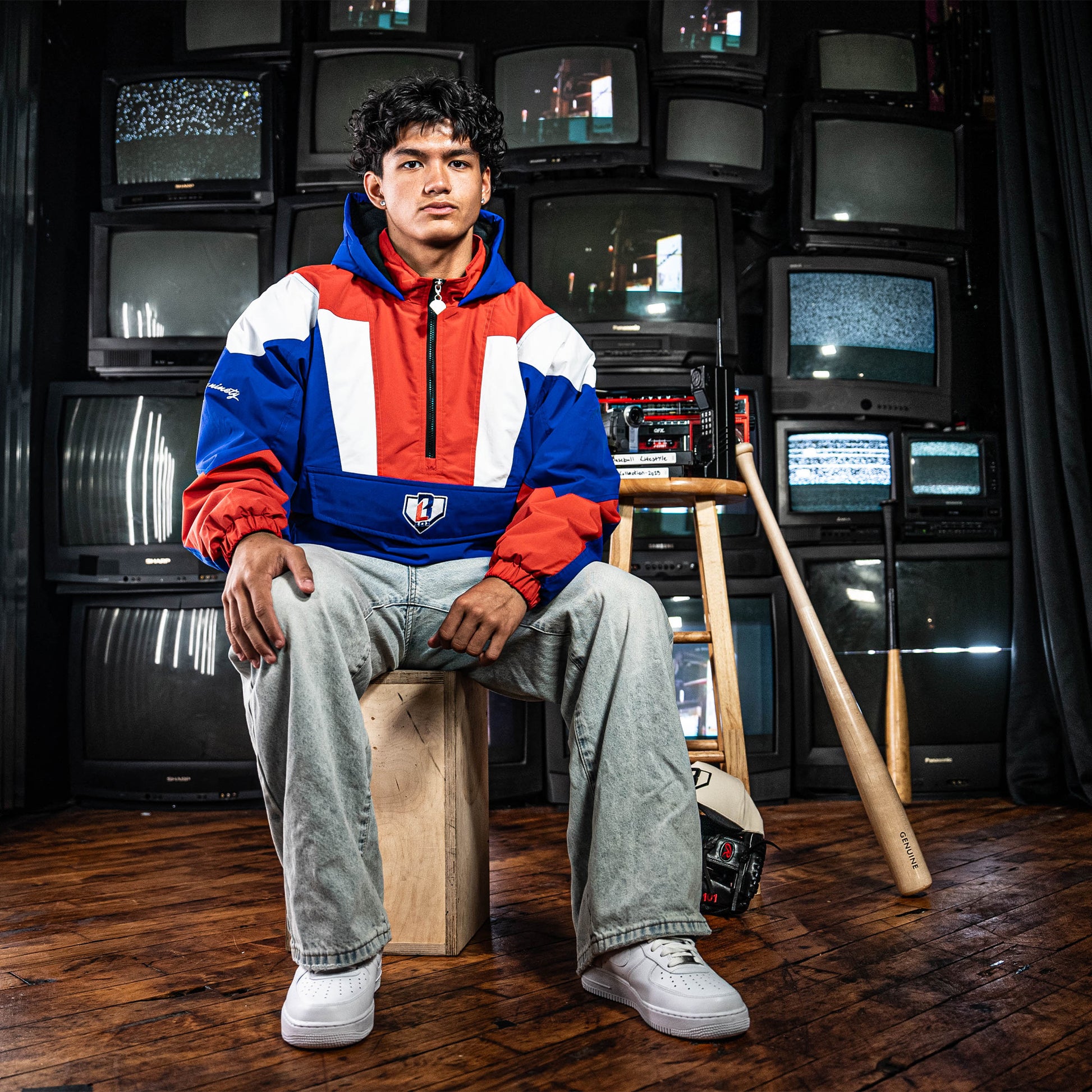 Person wearing a red, white, and blue jacket sitting on a wooden stool in front of vintage TVs.