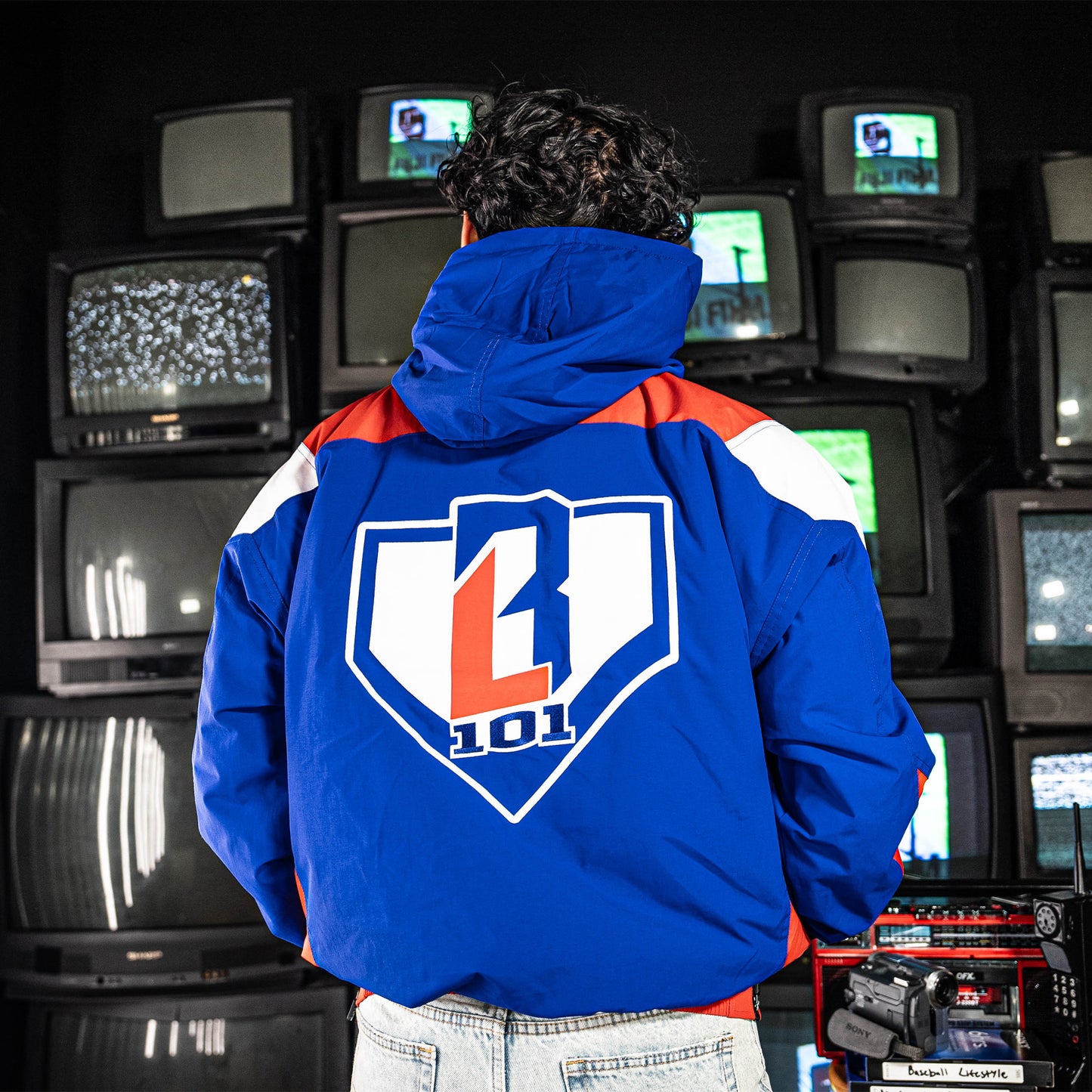 Person wearing a blue, red and white jacket with BL101 logo in front of vintage TVs.