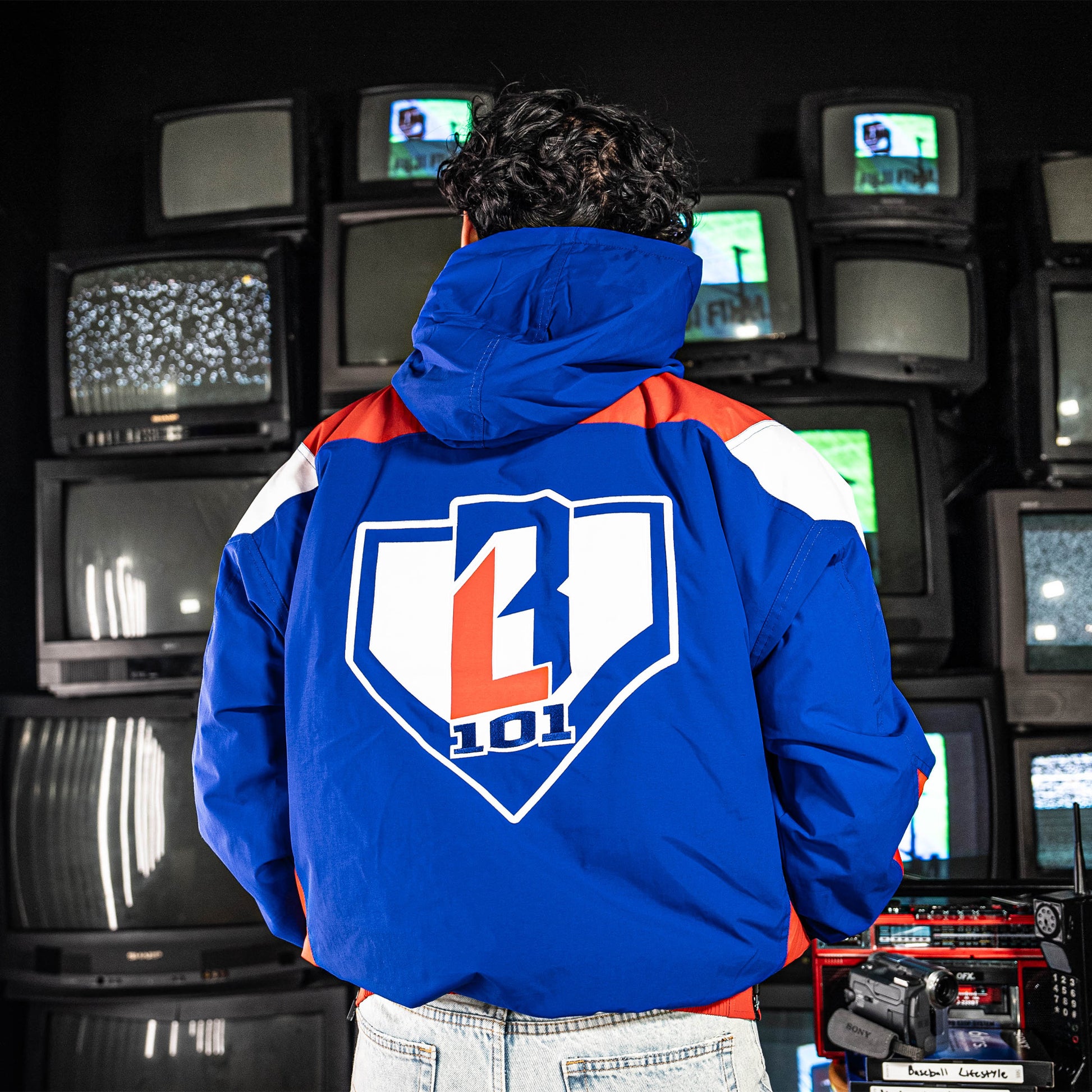 Person wearing a blue, red and white jacket with BL101 logo in front of vintage TVs.