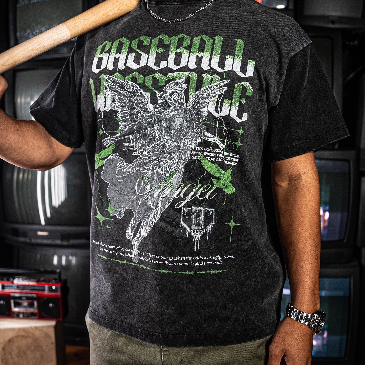 Person wearing a grey t-shirt with a green and white graphic design and text, holding a baseball bat.
