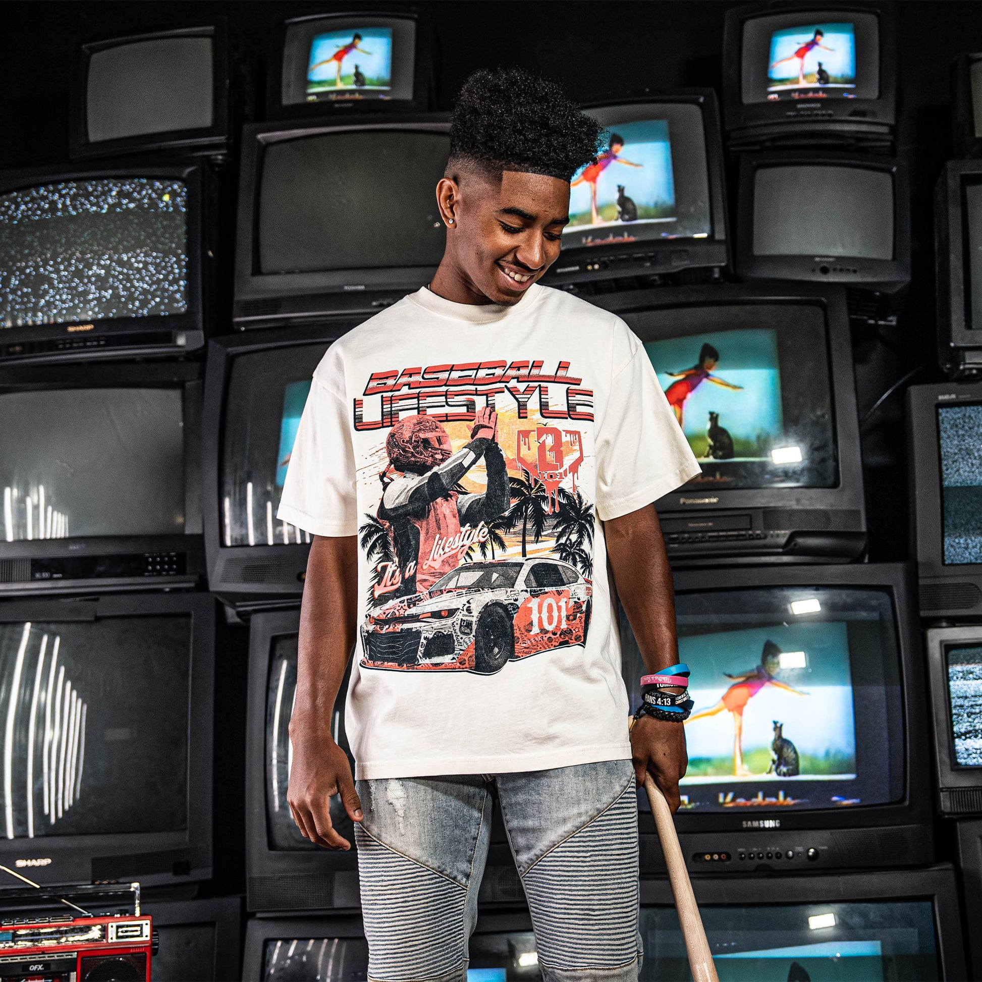 Person wearing a white t-shirt with a graphic design of a car and 'Baseball Lifestyle' text, standing in a room with stacked tv's.