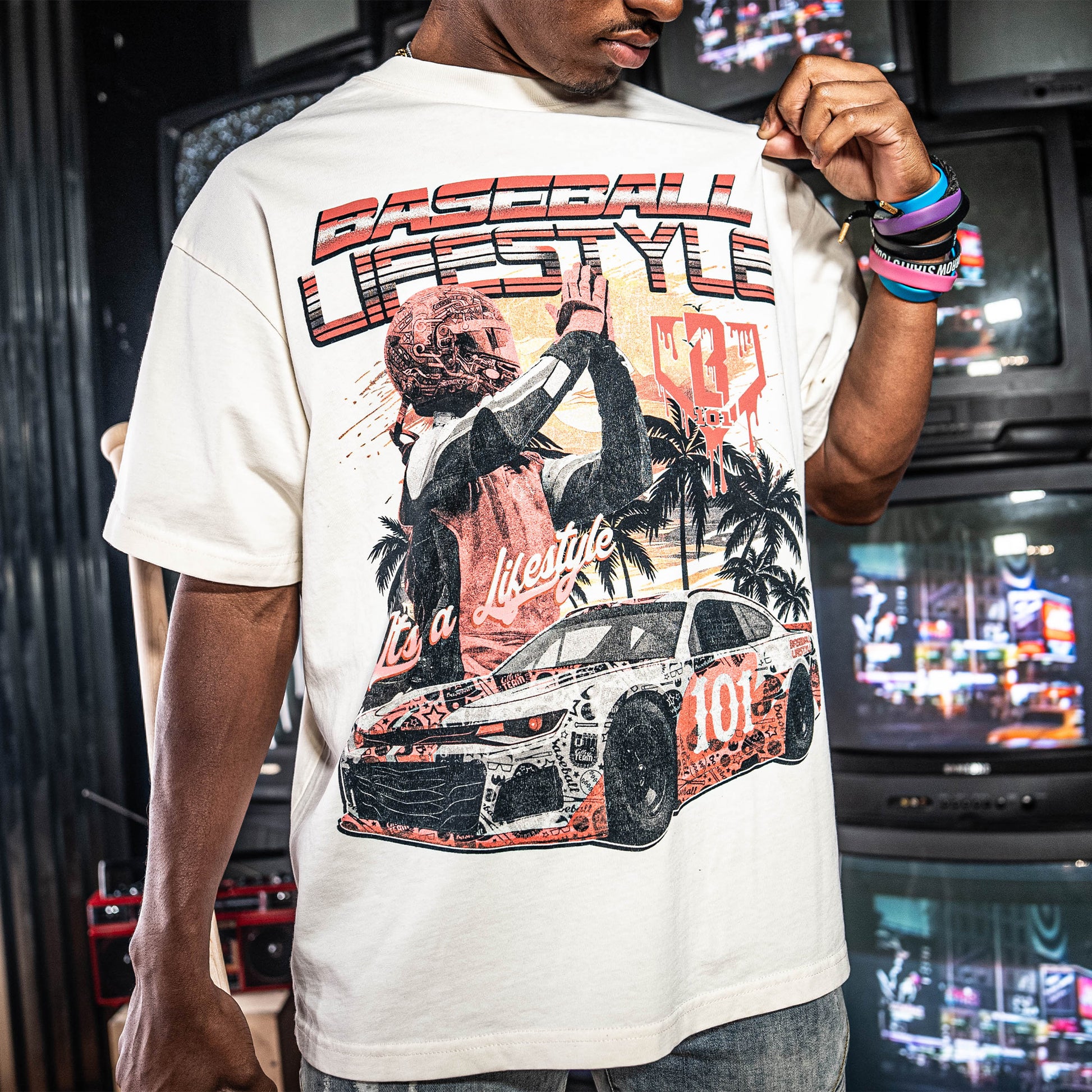 Person wearing a white t-shirt with a graphic design of a car and 'Baseball Lifestyle' text, standing in a room with stacked tv's.