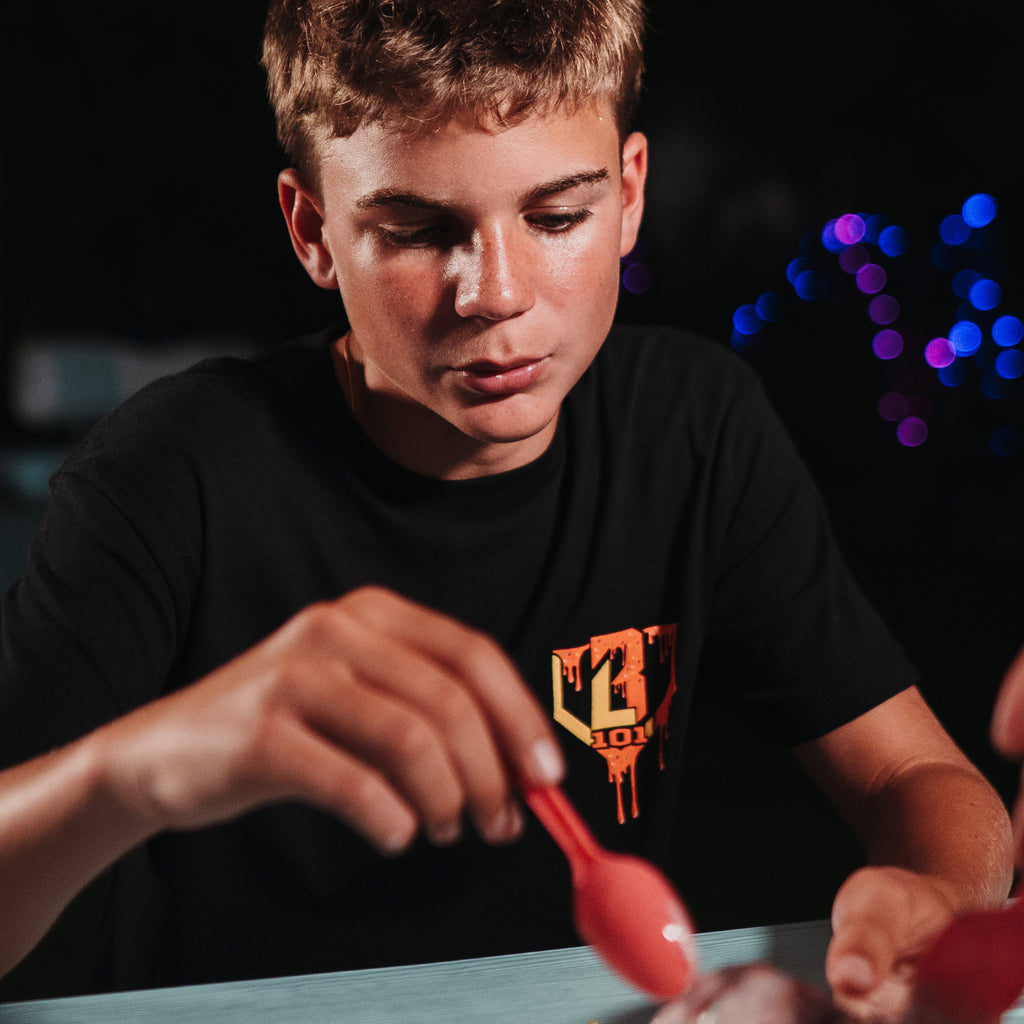 Person wearing a black t-shirt with a red and yellow BL101 logo, holding a red object against a dark background with colorful lights.