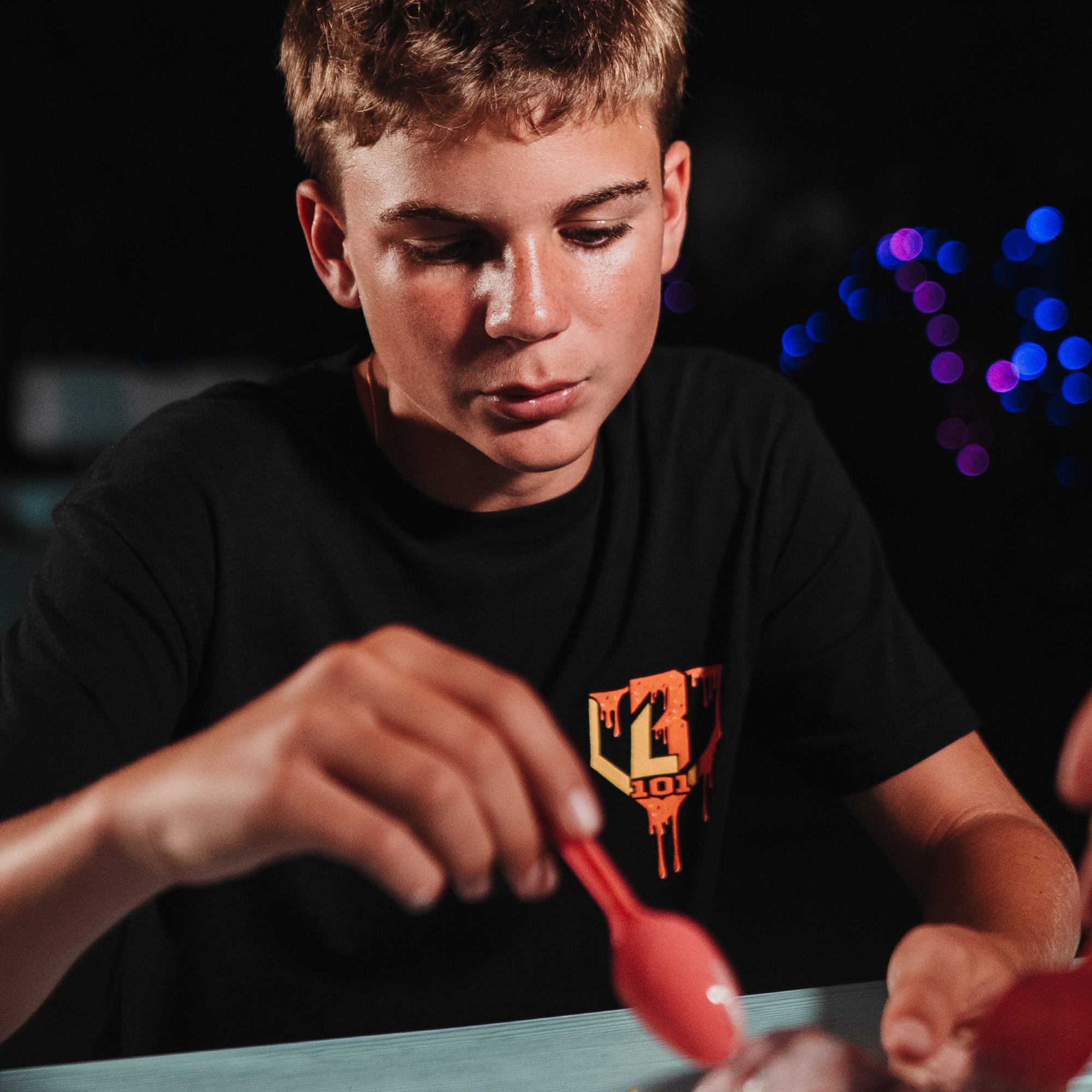 Person wearing a black t-shirt with a red and yellow BL101 logo, holding a red object against a dark background with colorful lights.