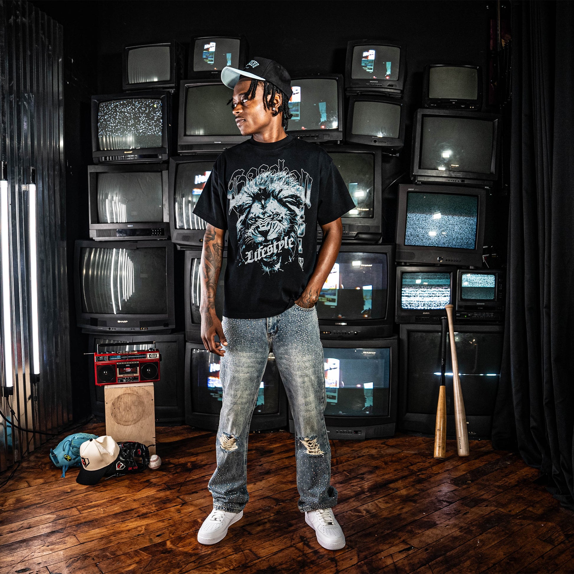 Person wearing a Black t-shirt with blue lion graphic and 'Baseball Lifestyle' text standing 9in front of TV's