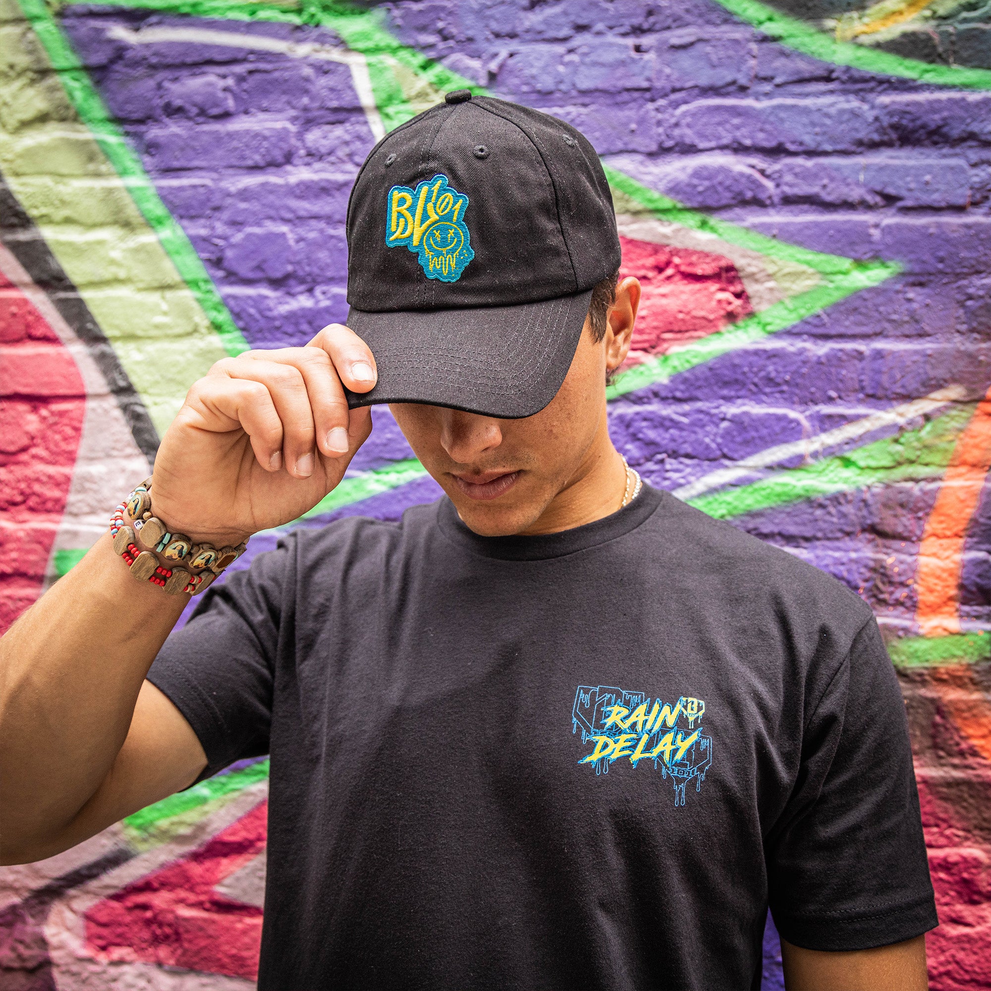 Person wearing a black cap and t-shirt with colorful graphics against a graffiti-covered wall.