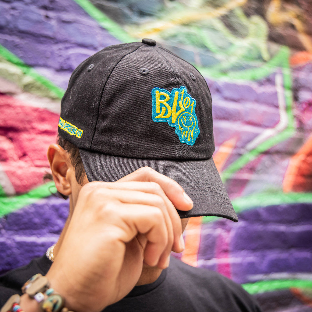Person wearing a black cap with blue and yellow BL101 logo and smiley face against a graffiti wall