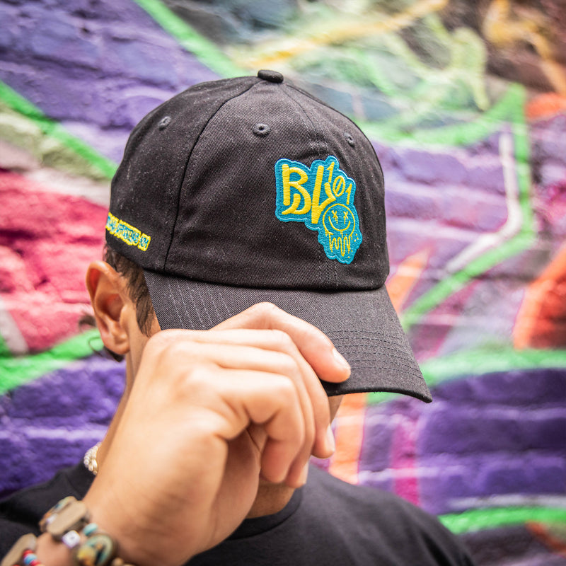 Person wearing a black cap with blue and yellow BL101 logo and smiley face against a graffiti wall