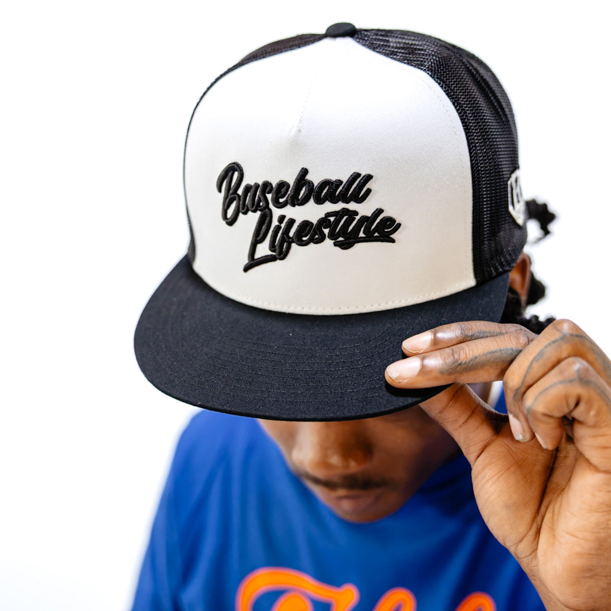 Person wearing a black and white cap with 'Baseball Lifestyle' text, holding it with one hand.