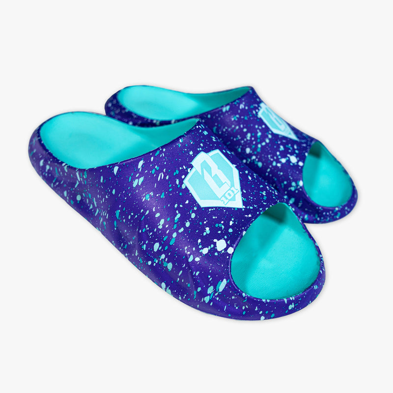 Person wearing BL101 purple splatter slides and matching swim trunks poolside, holding a matching purple splatter towel.