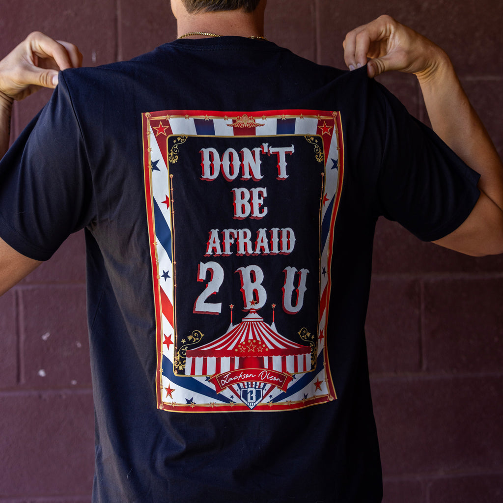Person wearing black t-shirt with a graphic design featuring 'Don't Be Afraid 2 B U' text and a circus tent