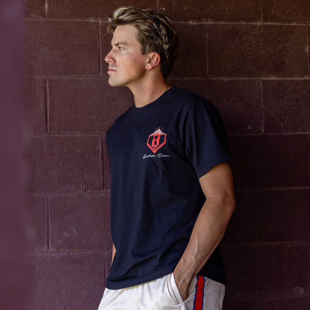 Man wearing black t-shirt with a small red and white 'BL101 Jackson Olson' logo on the chest against brick wall 